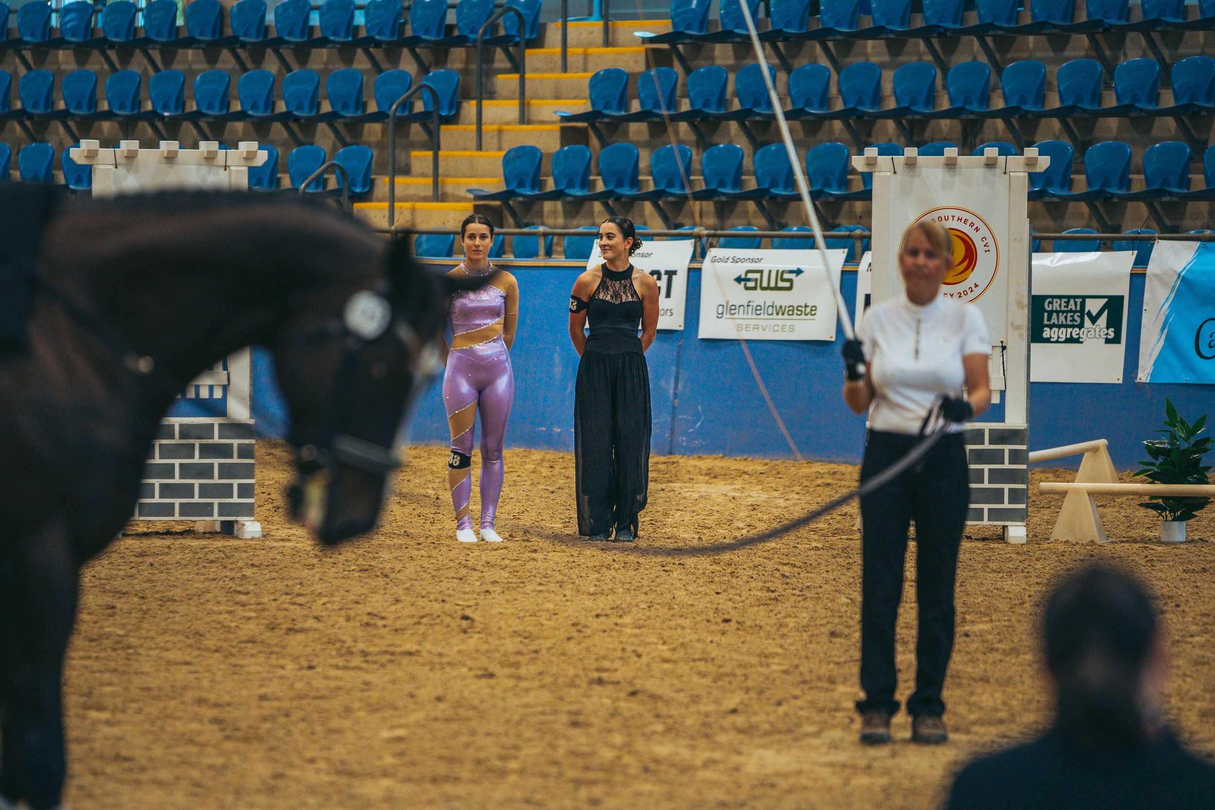 A woman leading a horse during an indoor equestrian event, with two women standing in the background and audience seating visible.