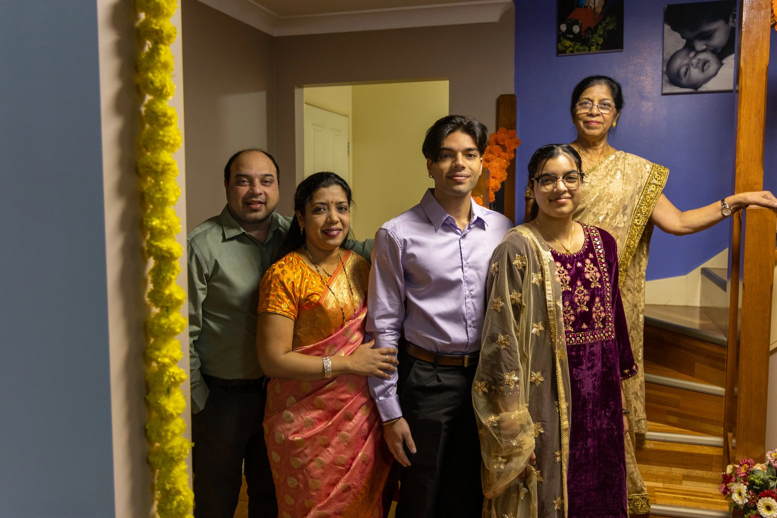 Family celebrating indoors, standing on stairs, four women, two men, some wearing traditional Indian attire, decorated for a special occasion.