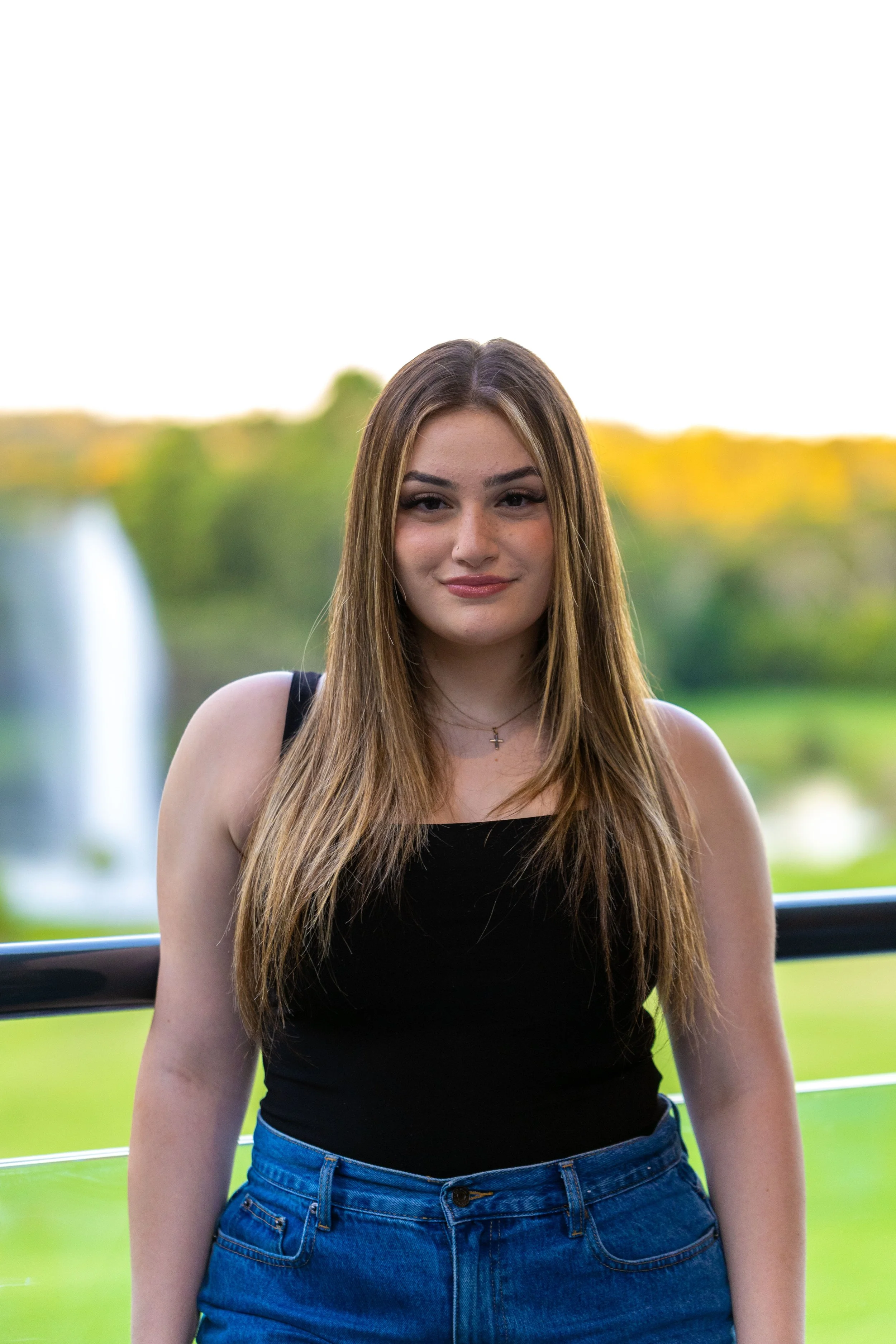 A young woman with long brown hair, wearing a black tank top and blue jeans, standing outdoors with a blurred background of greenery and a water fountain.
