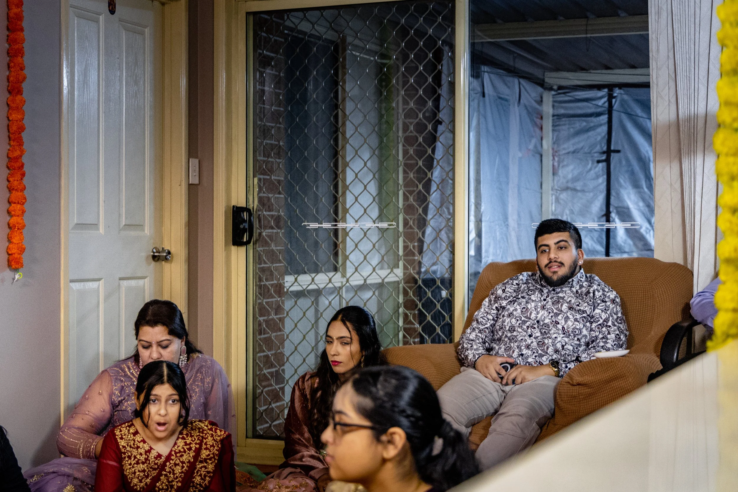 A group of people sitting indoors, three women and one man visible, with some of the women wearing traditional South Asian attire, in a room decorated with marigold flower strings.