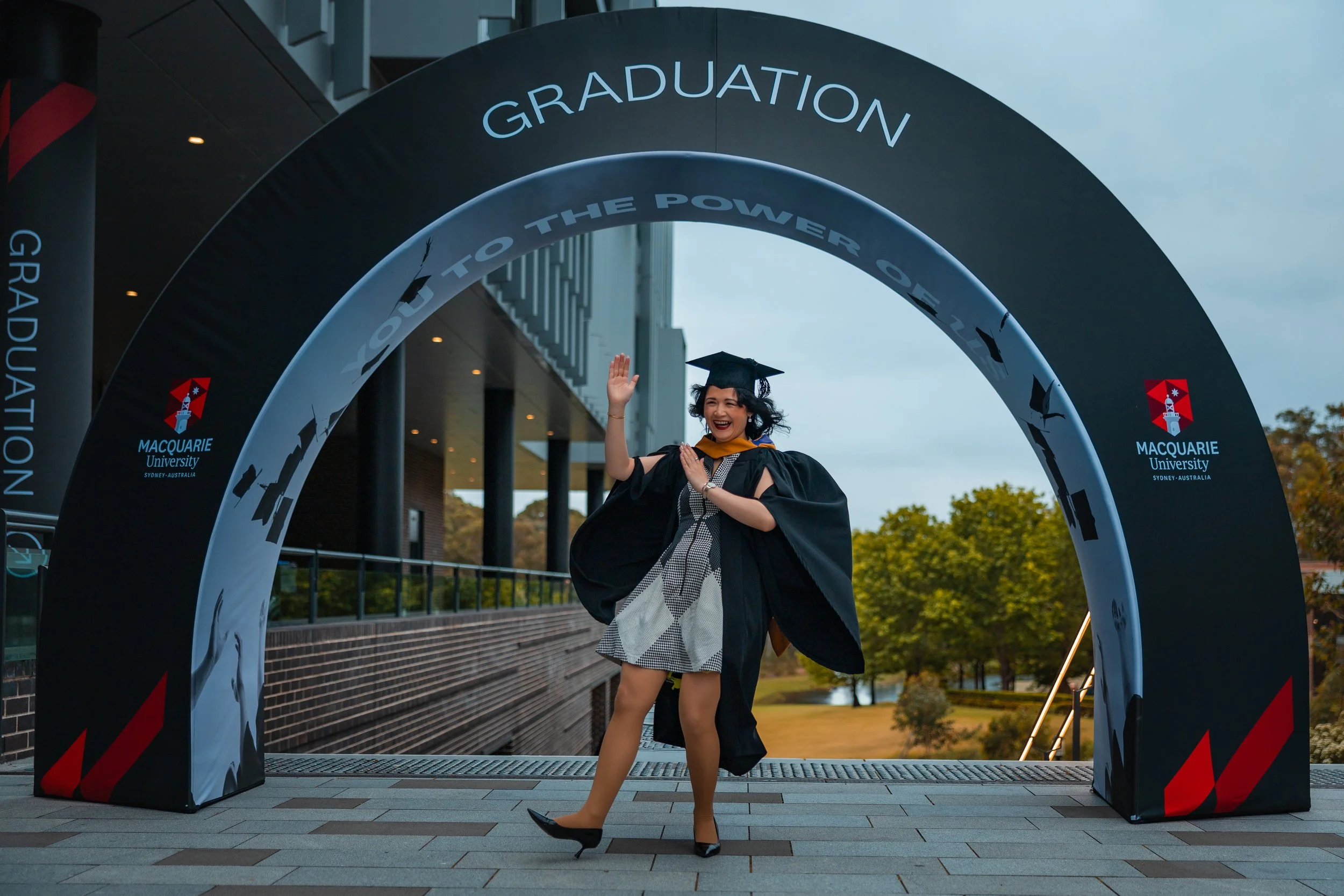 A woman celebrating her graduation in front of a Macquarie University archway, wearing a black gown and mortarboard, smiling, with trees and a building in the background.