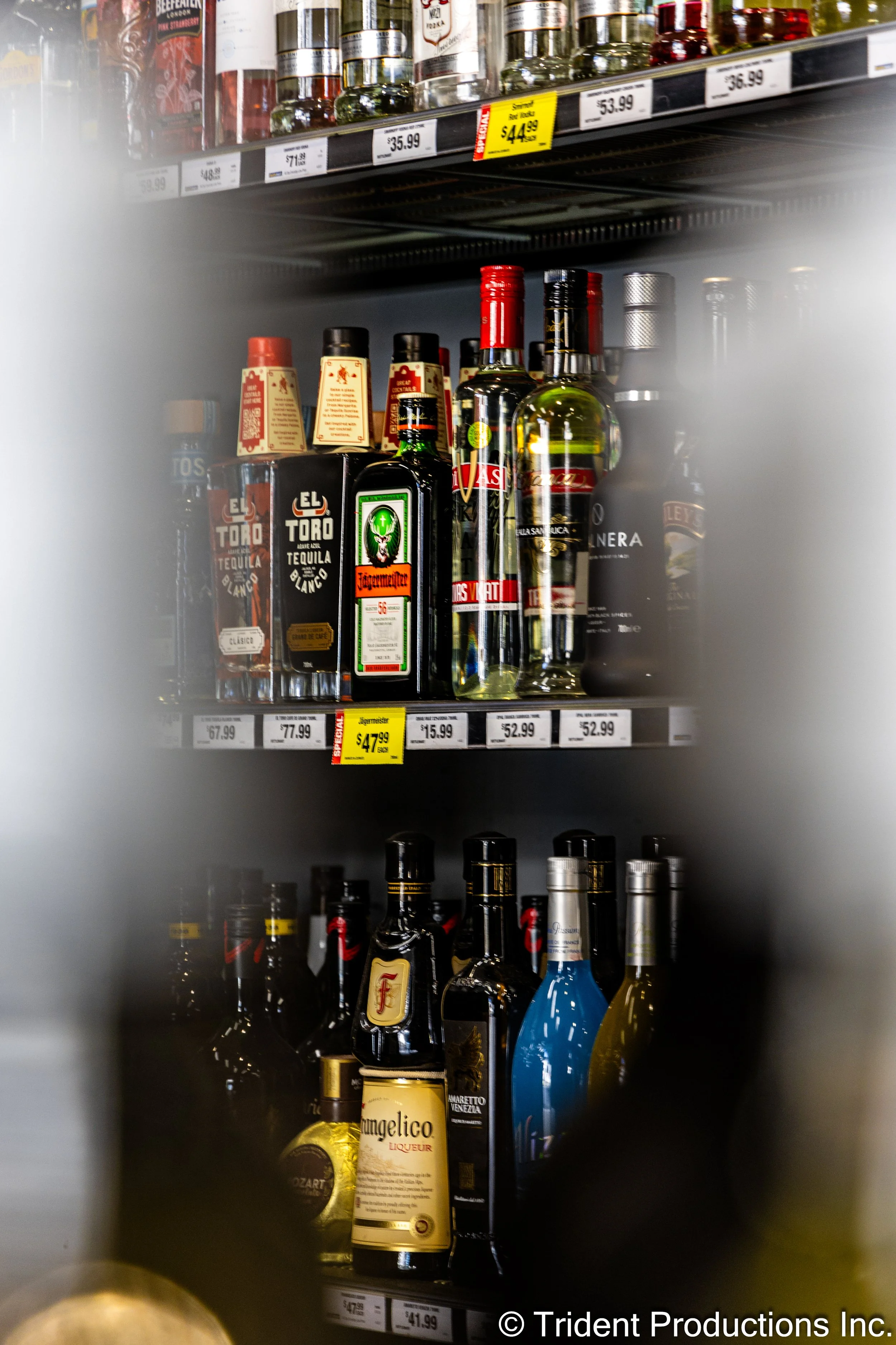 Assorted bottles of liquor behind a grocery store shelf, with some price tags visible, including tequila and flavored liqueurs, partially obscured by a blurred foreground.