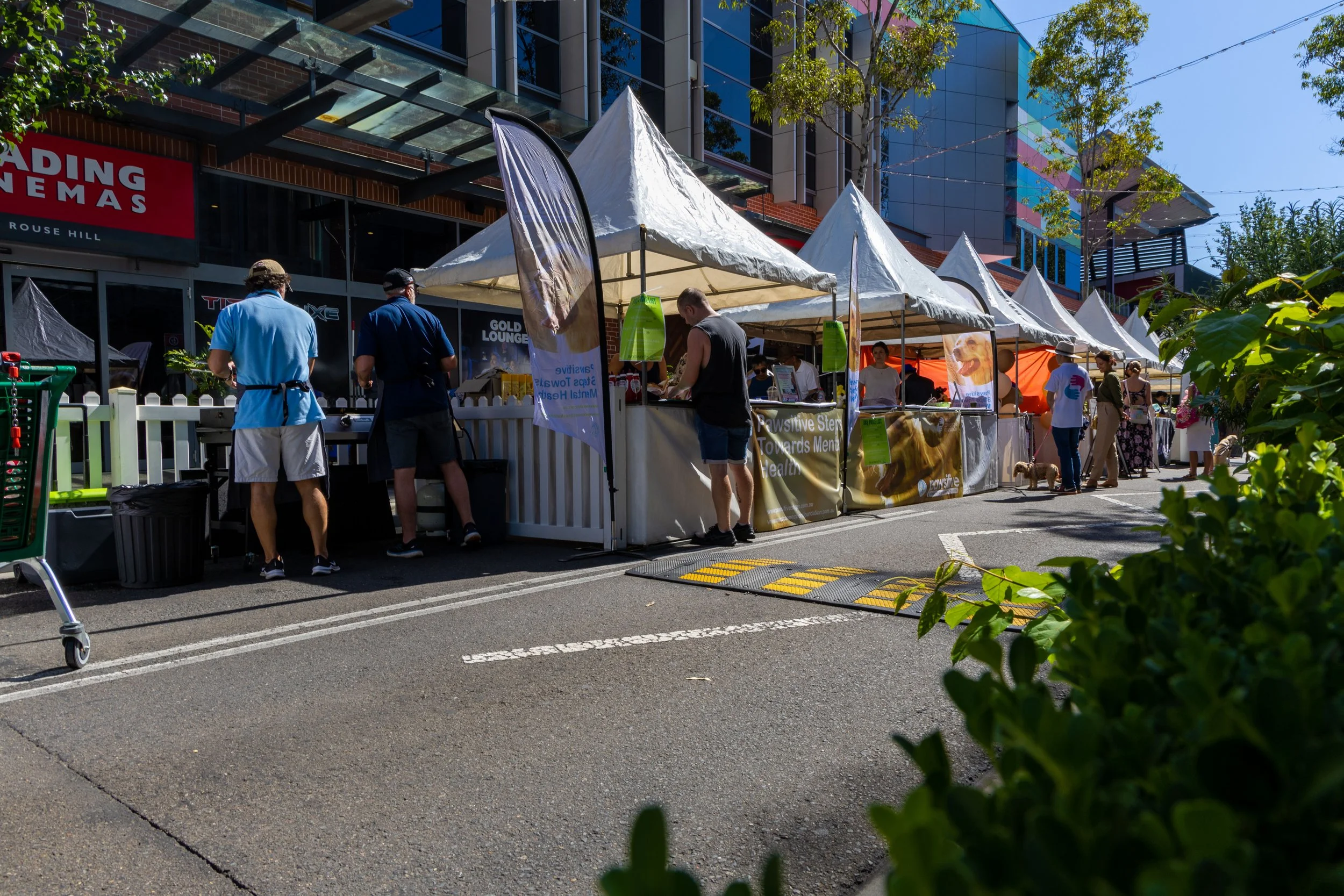 Outdoor market with white tents and people browsing and shopping, situated on a city street with modern buildings in the background.