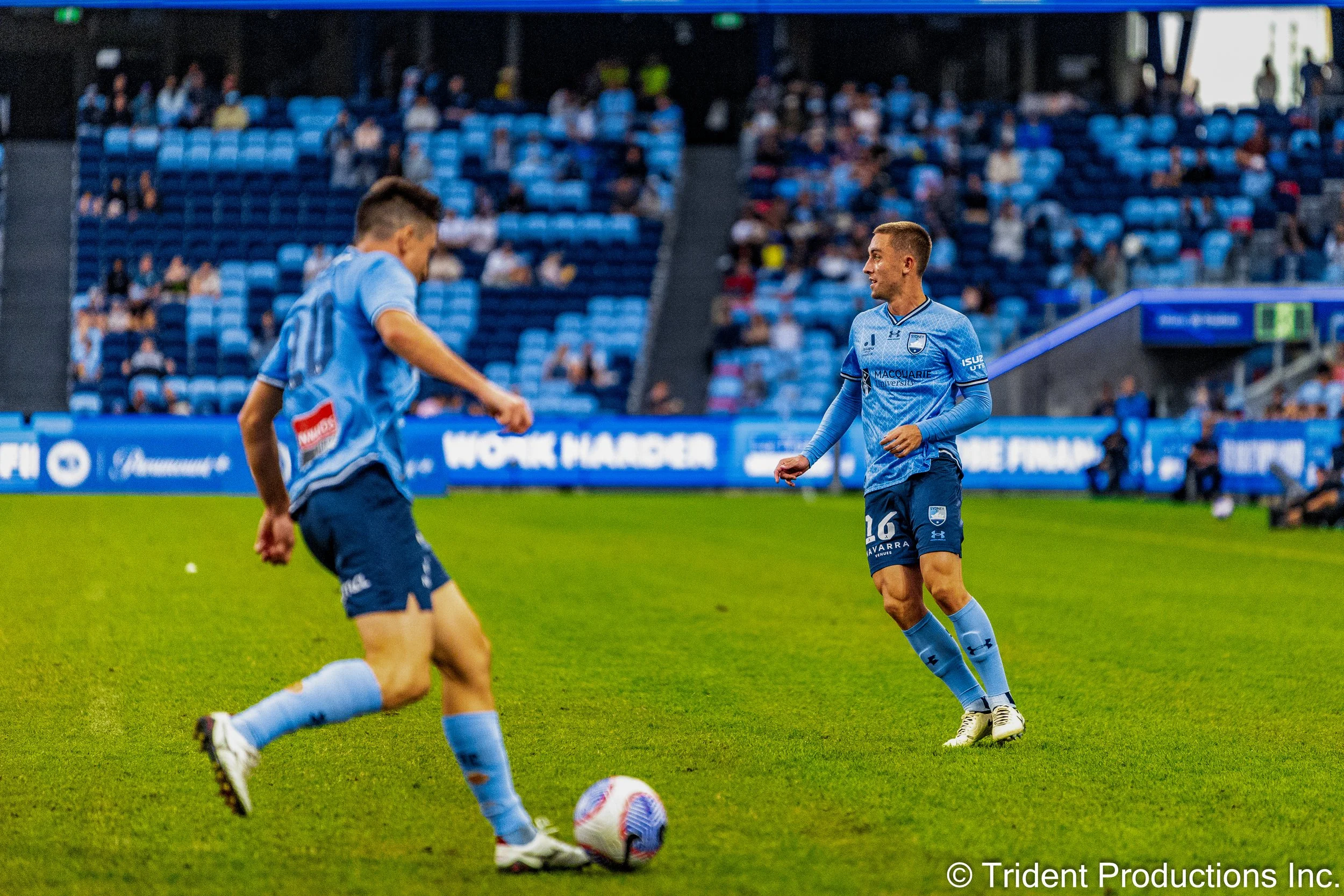 Two soccer players in blue uniforms warming up on the field, with a crowd in the stands behind them.