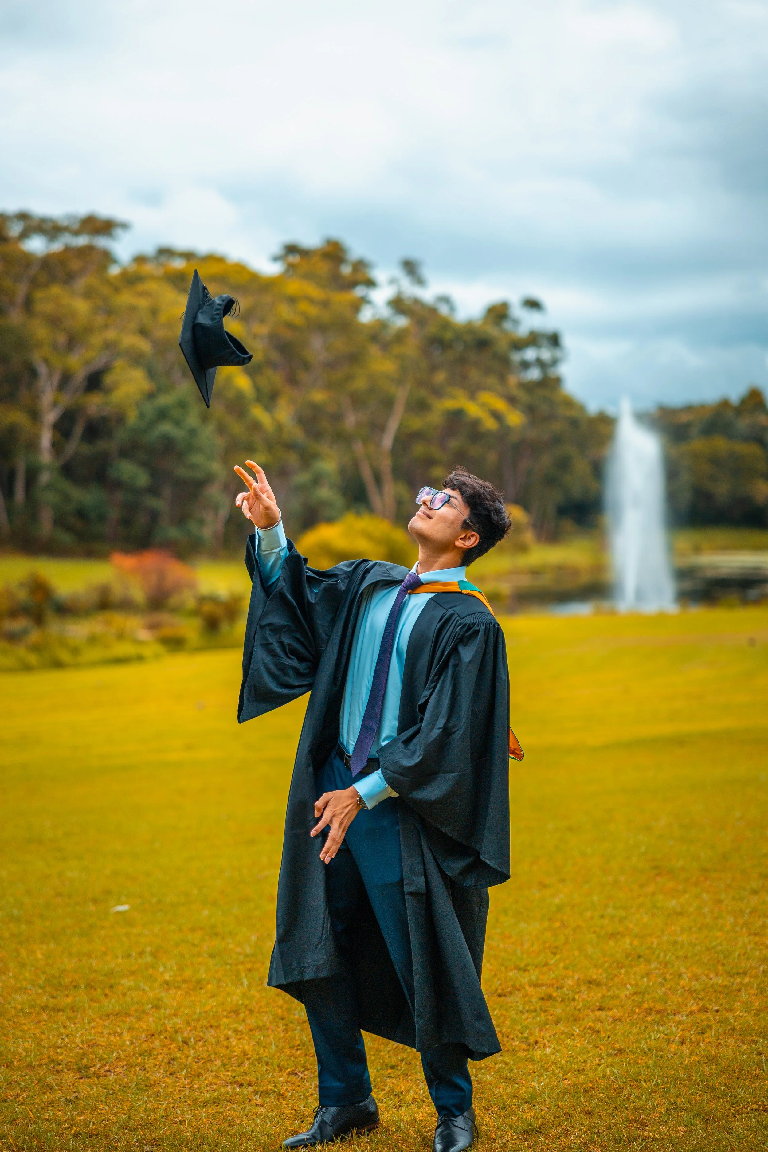 A young man in a graduation gown and cap throwing his cap into the air outdoors, with trees and a fountain in the background.
