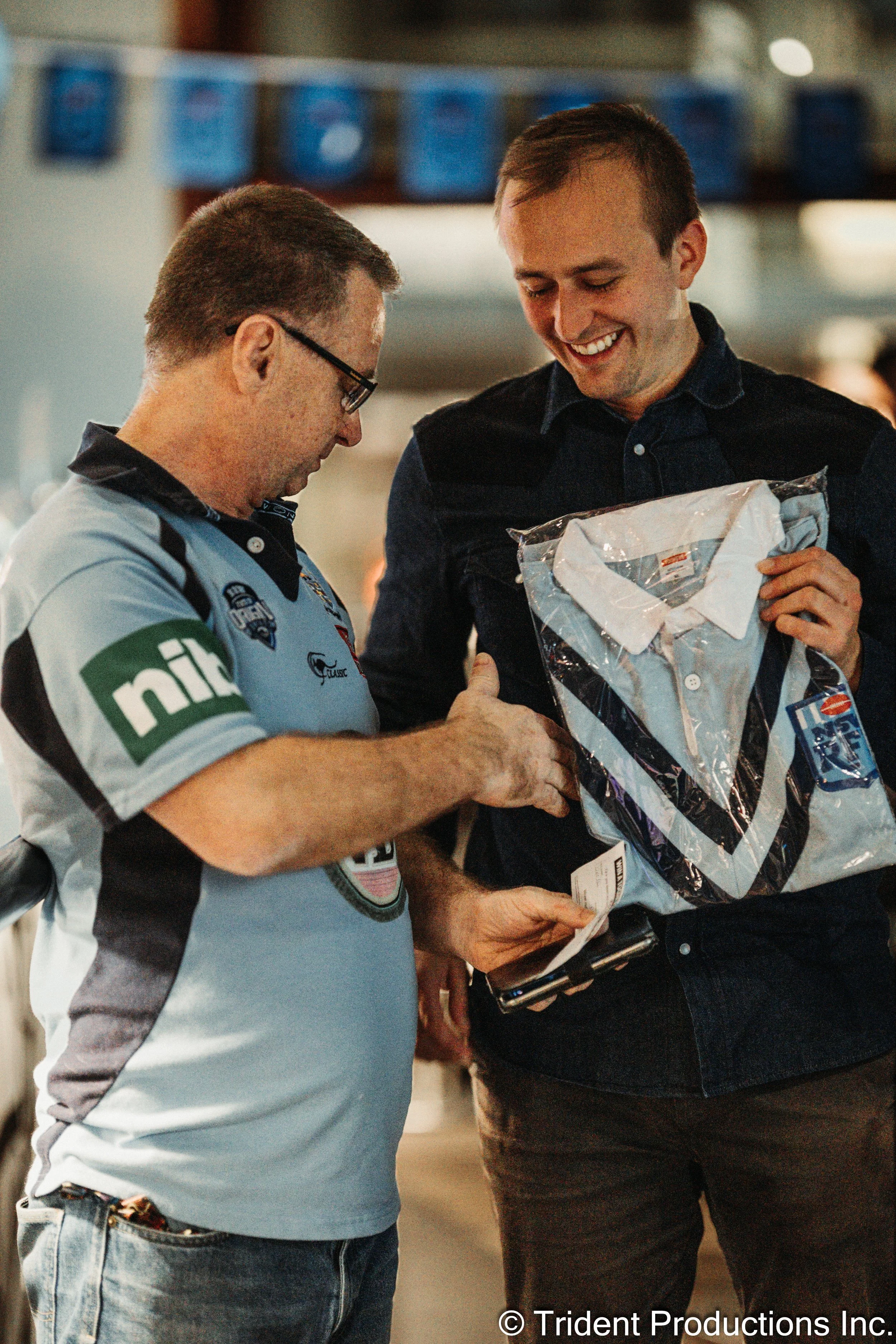 Two men at an event; one in a NASCAR shirt, the other in a black shirt, smiling as they exchange a gift package wrapped in clear plastic with a blue and white striped shirt inside.