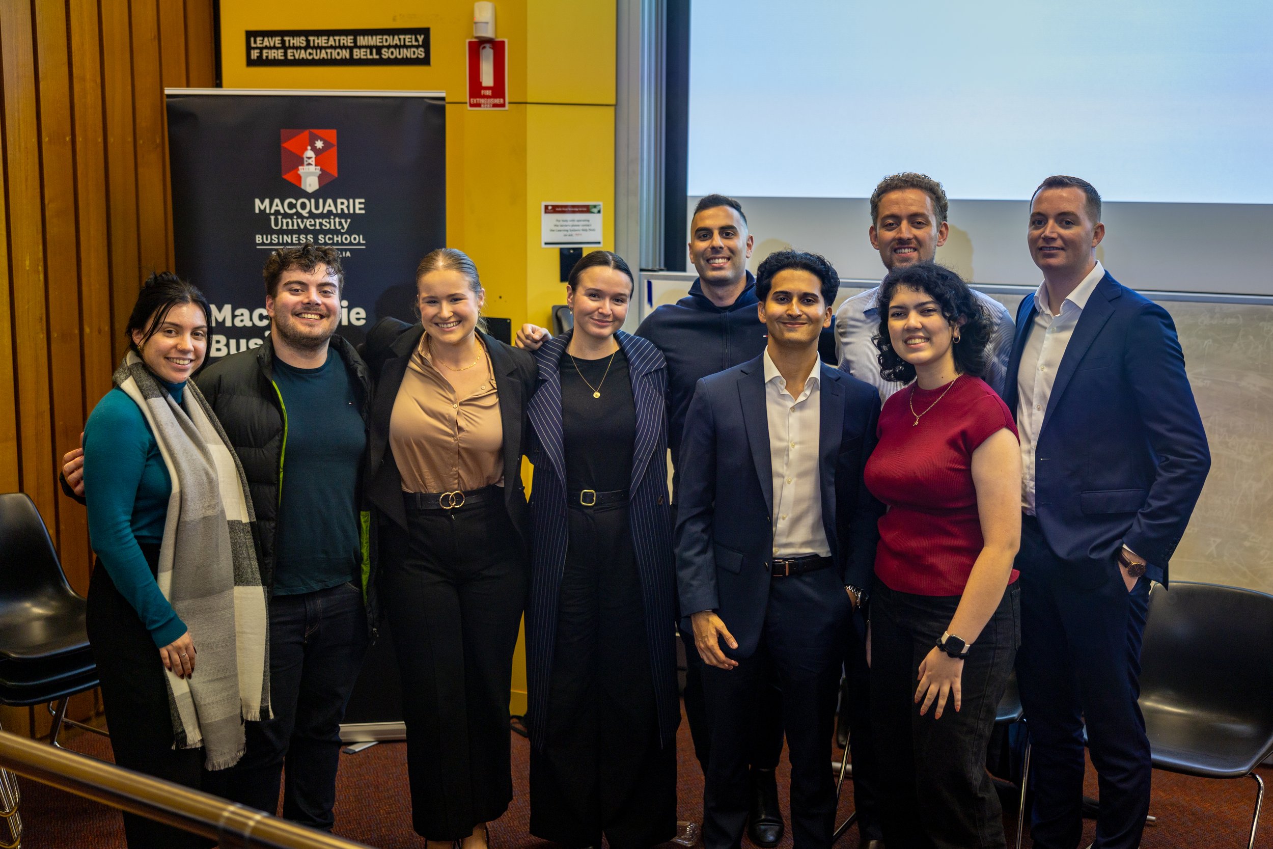 A group of nine diverse students standing together in a classroom or auditorium, smiling for a photo in front of a Macquarie University Business School banner and a projection screen.