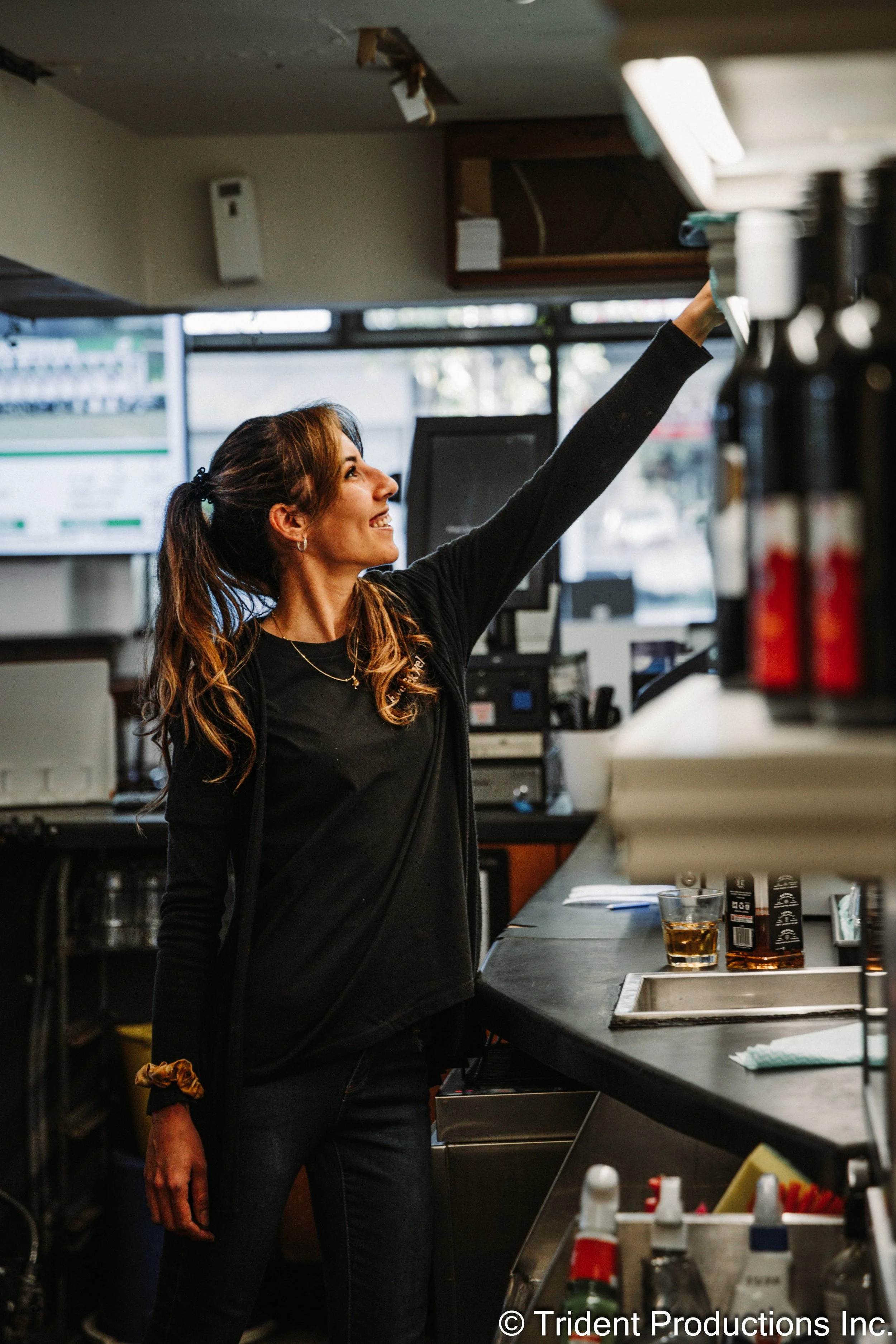 A woman working behind a bar counter, reaching up to grab an item from a shelf, with a drink on the counter and bar equipment around her.