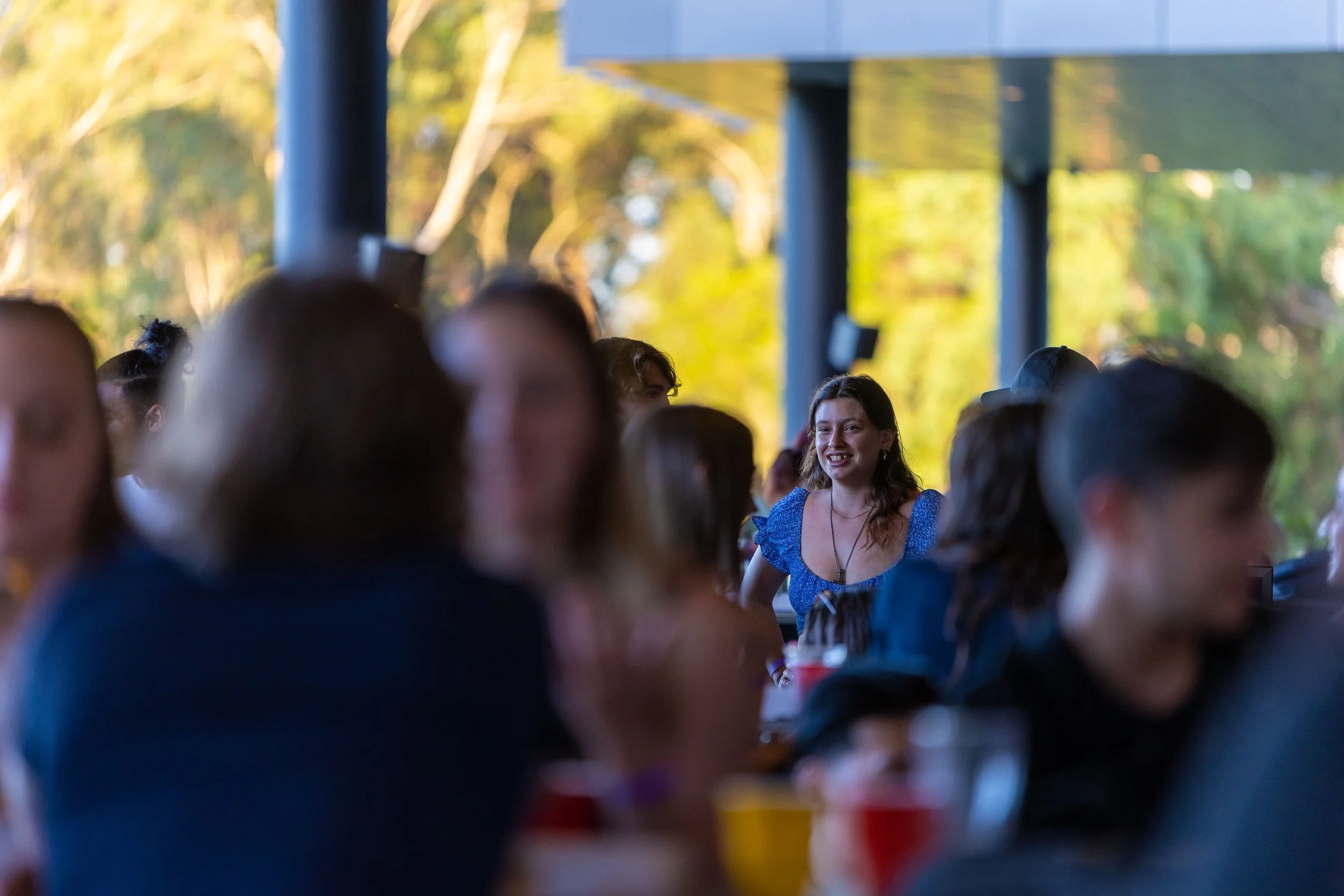 A group of people sitting at a social gathering or party, with a woman in a blue dress smiling and standing out in the background.