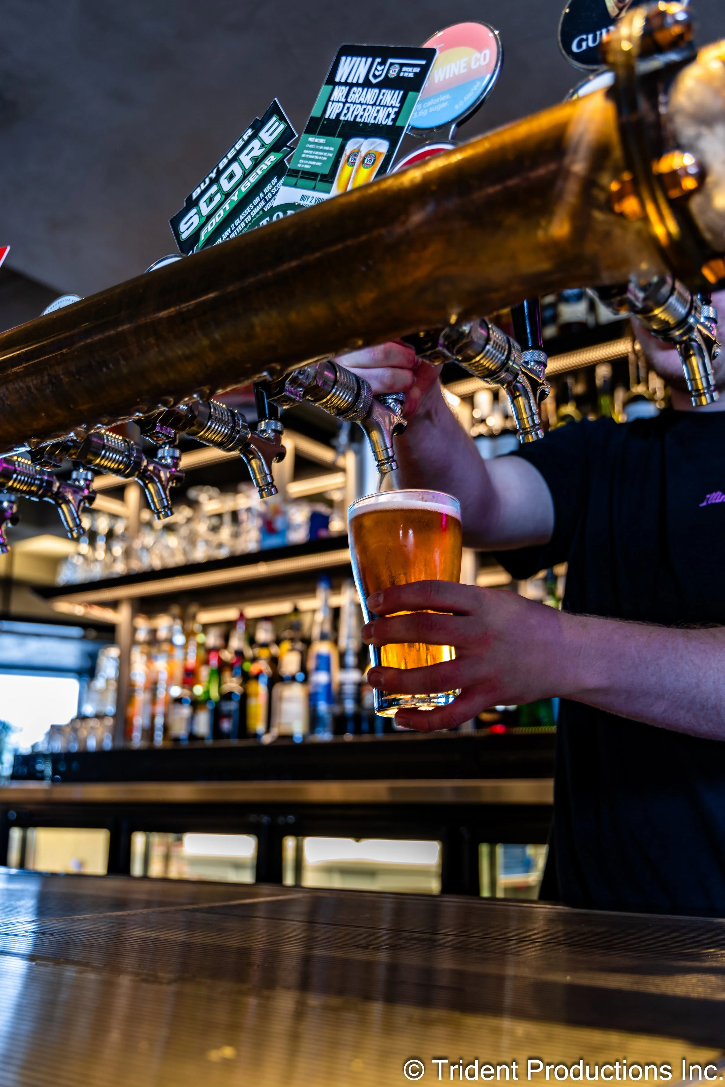 A bartender pours a beer from a tap into a glass at a bar with various bottles in the background.