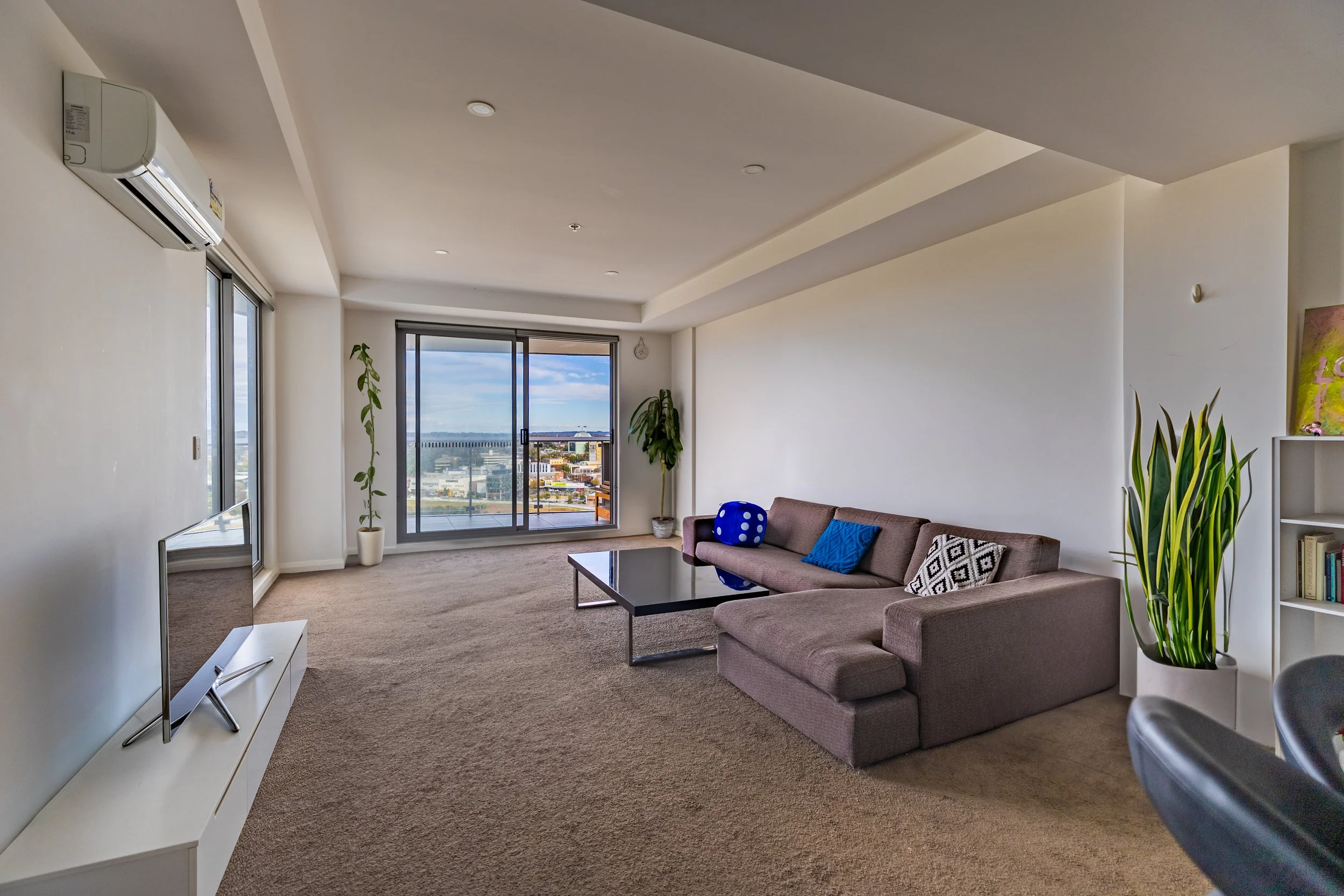 Modern living room with large sliding glass door overlooking cityscape, beige carpet, gray sectional sofa with patterned and solid blue throw pillows, black coffee table, potted plants, white bookshelf, wall-mounted air conditioner, and a flat-screen