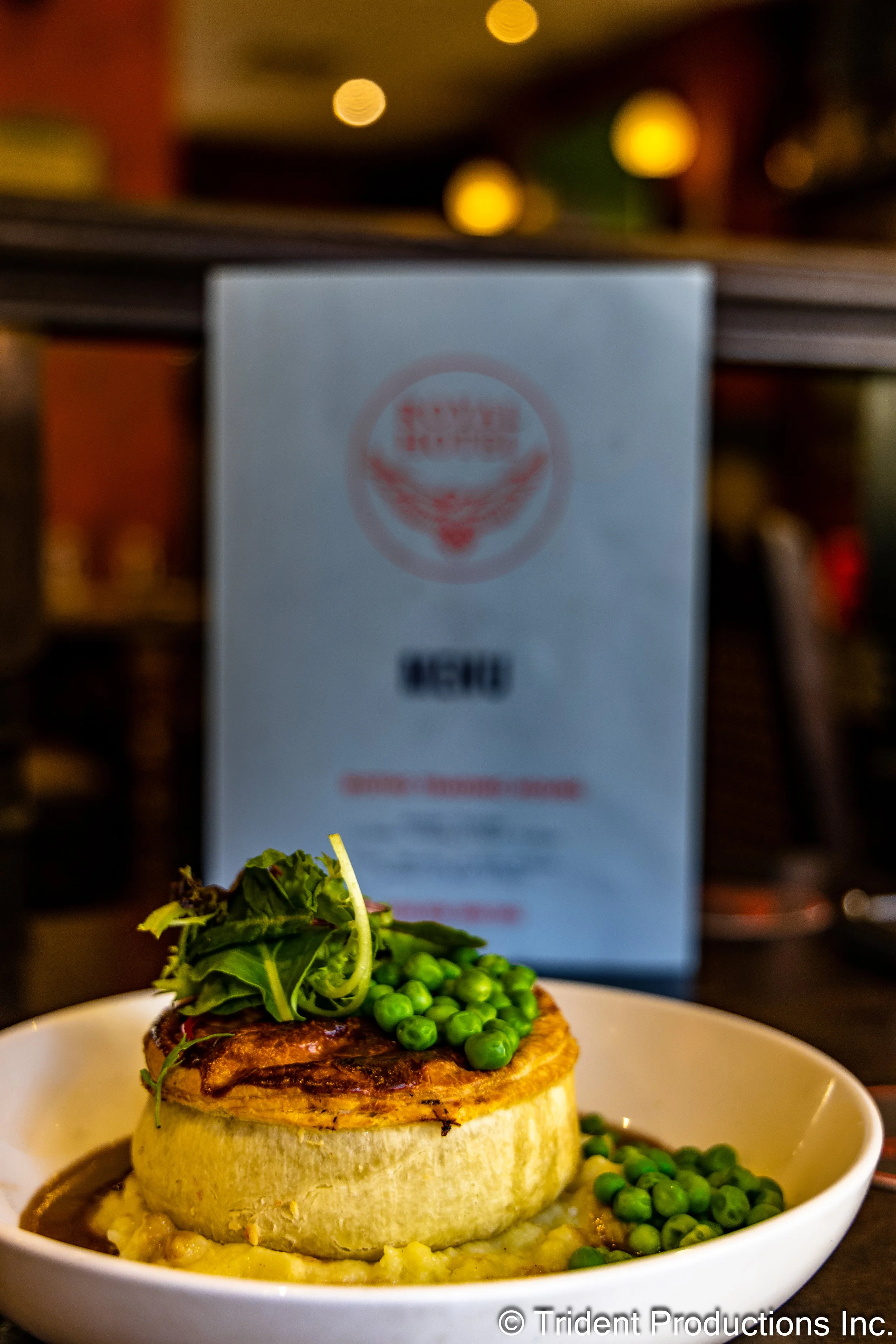 A plated gourmet burger on a white dish, topped with greens and green peas, with a blurry menu in the background.