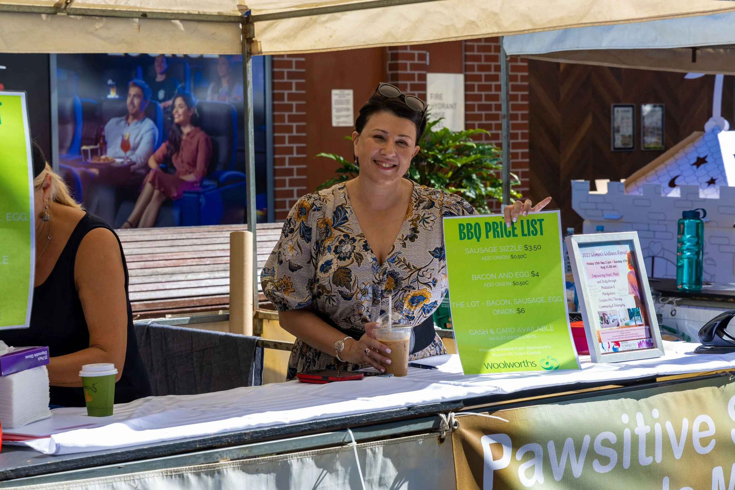 Woman smiling at a food stall holding a drink, with barbecue menu signs, in an outdoor setting.