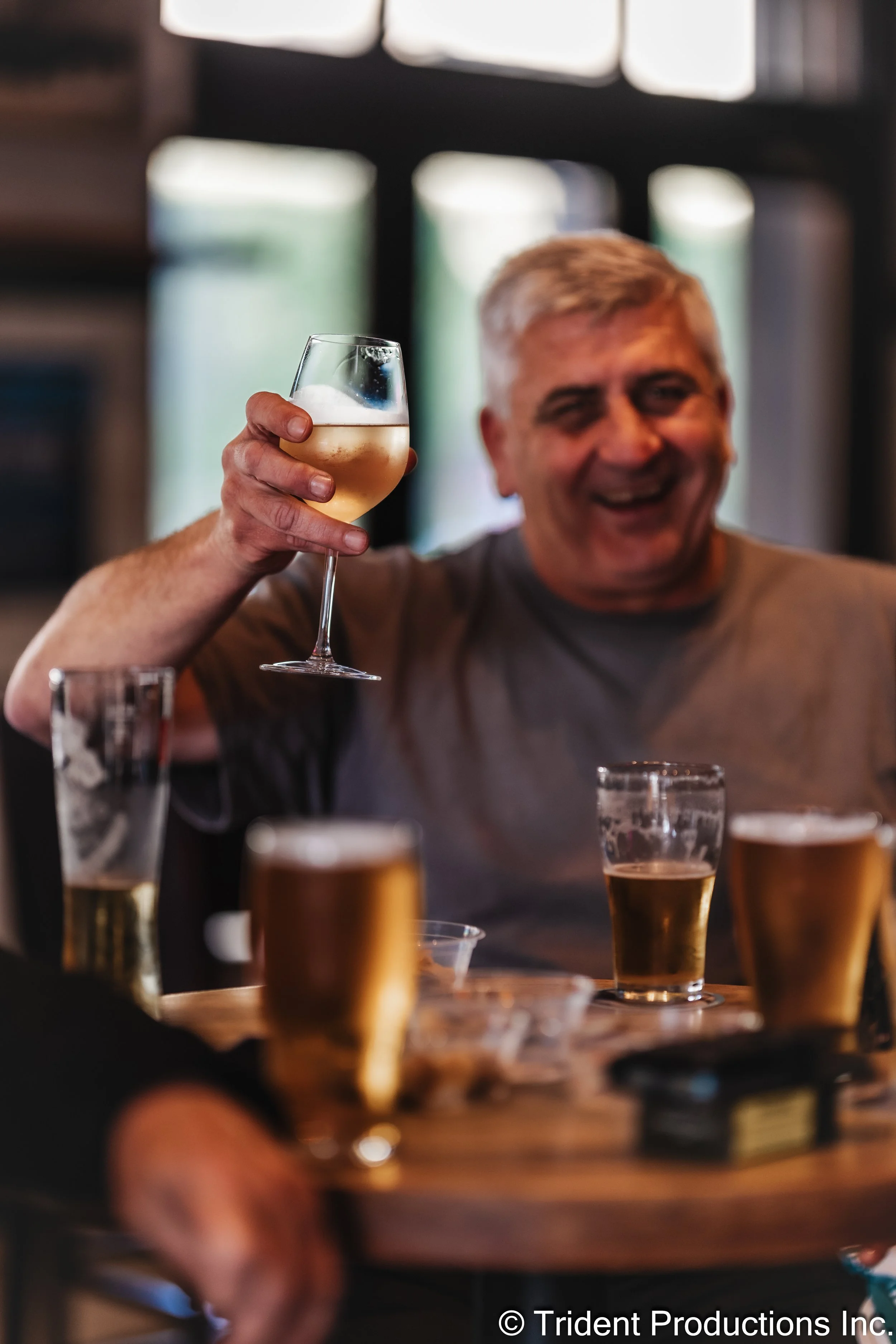 A smiling man with gray hair raising a glass of white wine in a toast. There are several other drinks on the table and a blurred background indoors.