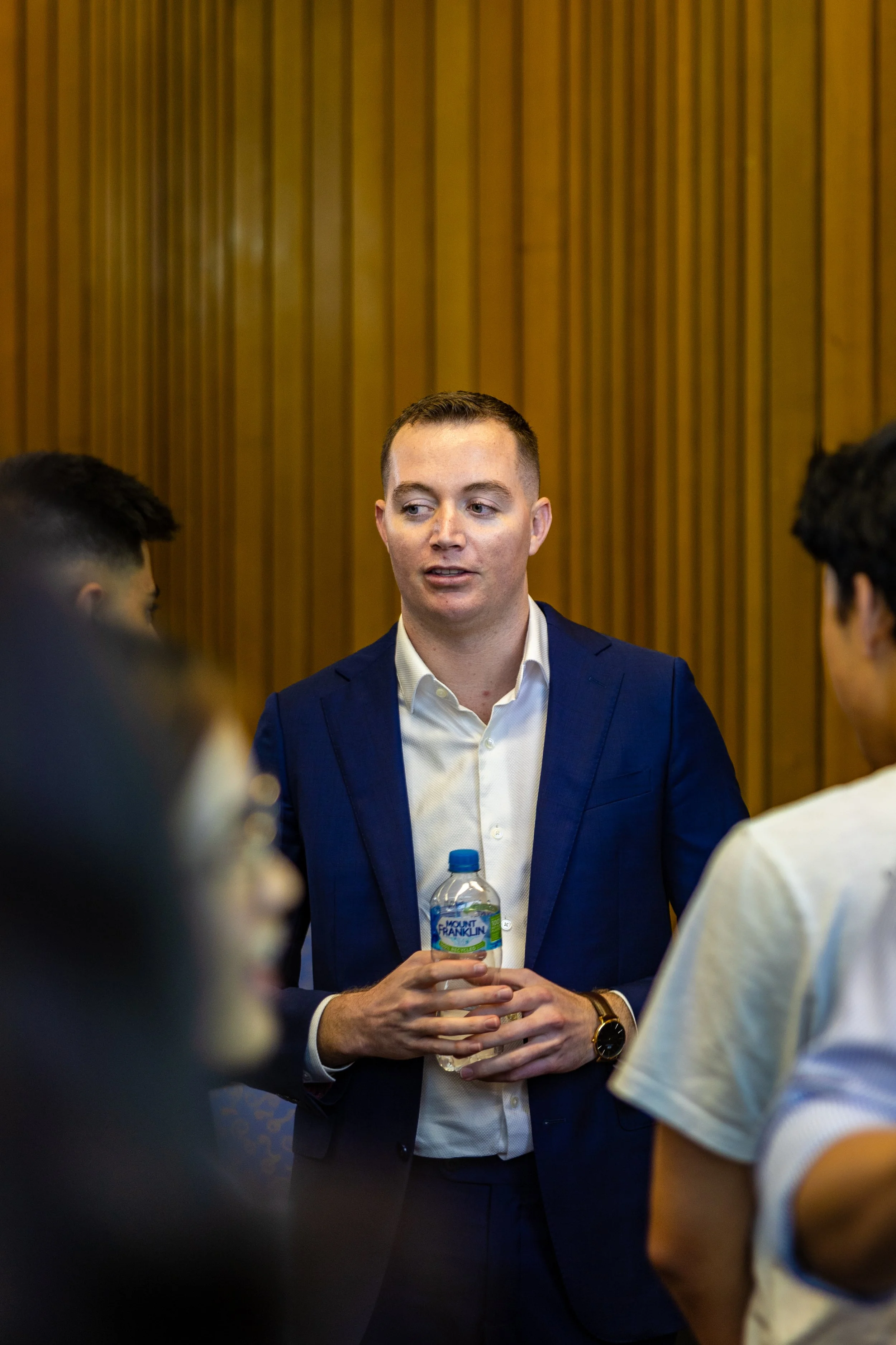 A man in a dark blue suit with a white shirt holding a water bottle, standing and talking with a group of people in a room with wooden paneling.