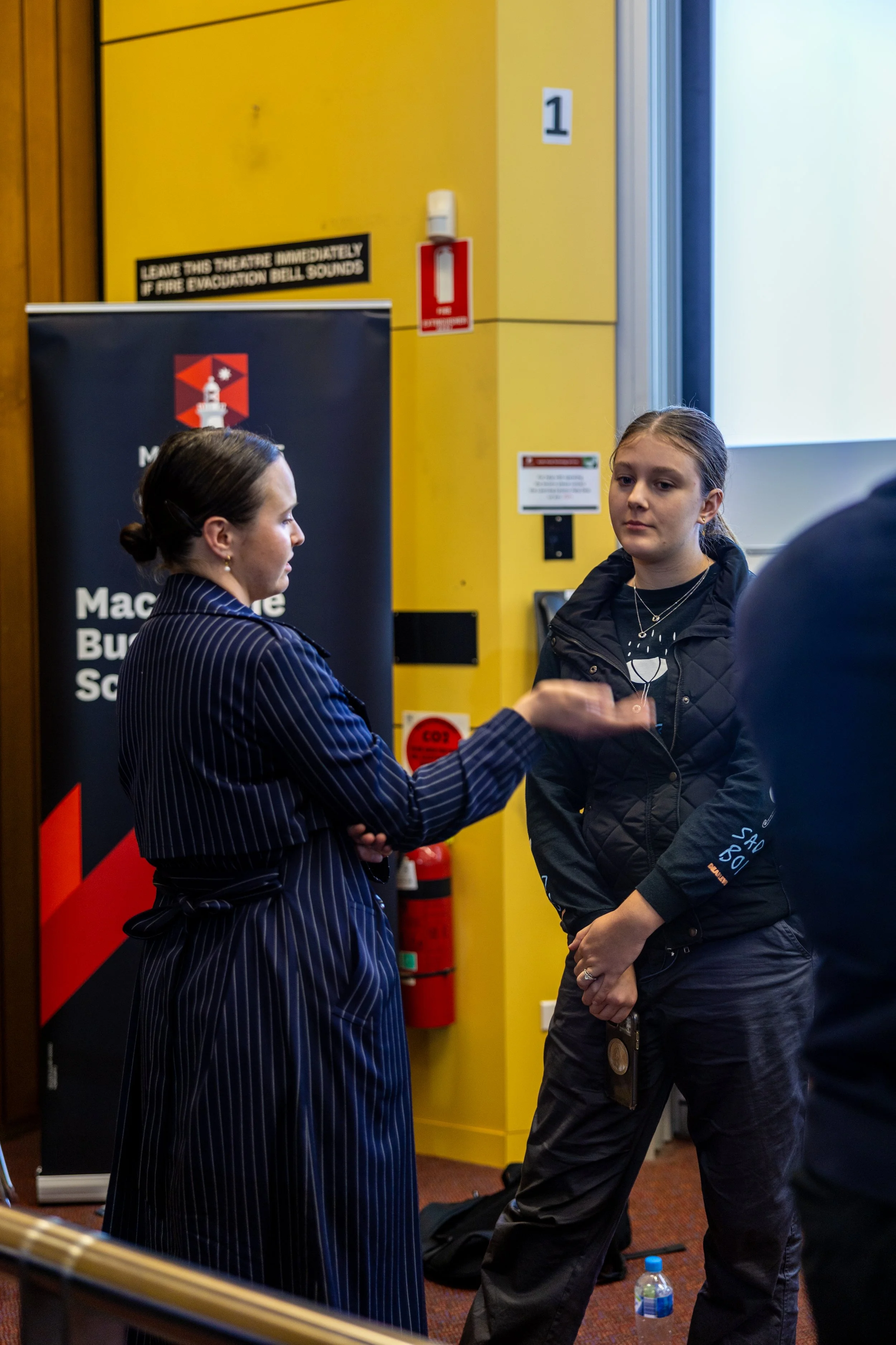 A woman in a striped blazer talks to a young girl in a black jacket inside a room with yellow walls, near a fire alarm and emergency instructions.