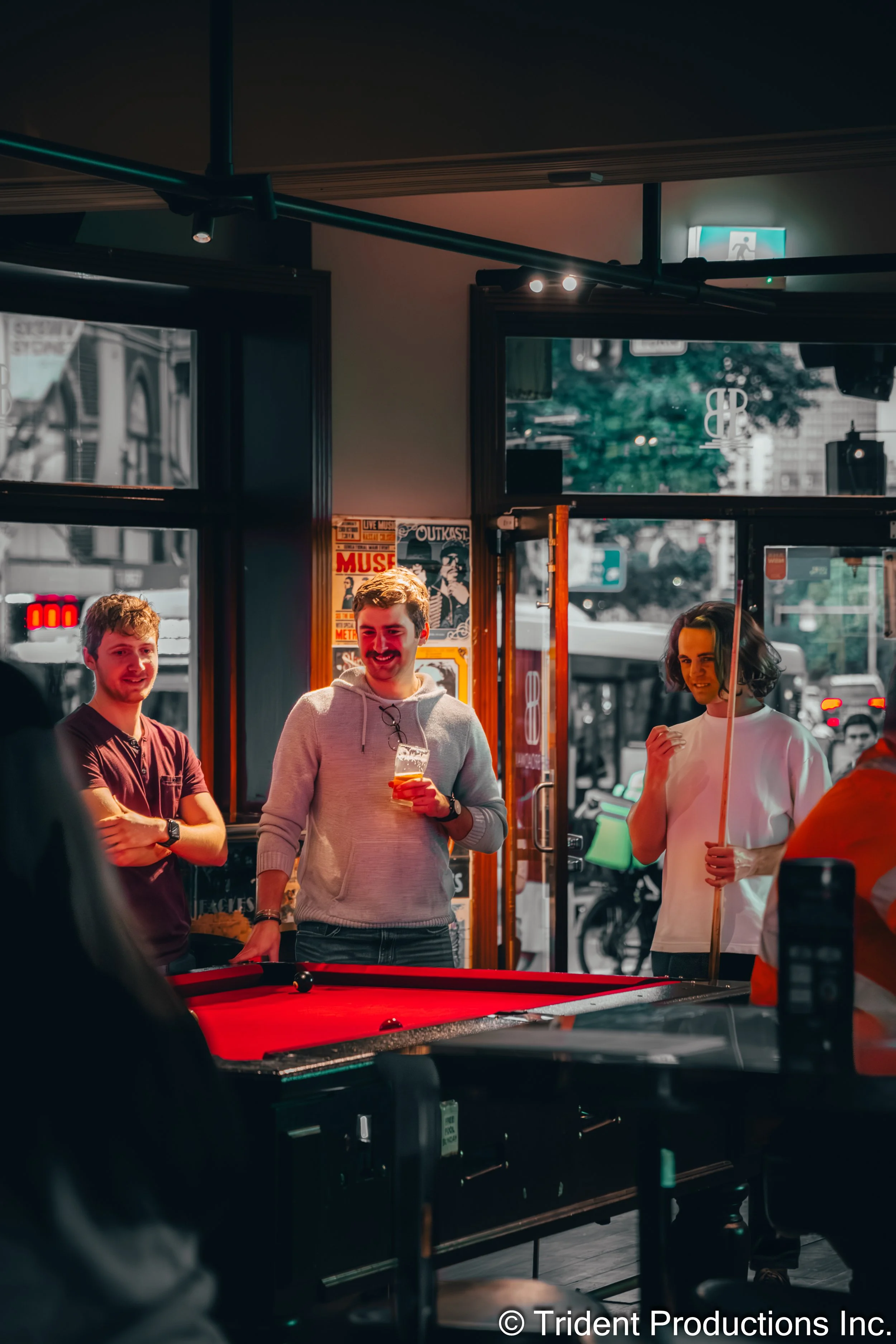 Three young adults stand near a pool table inside a bar, smiling and holding drinks. The bar has large windows with a city street visible outside.