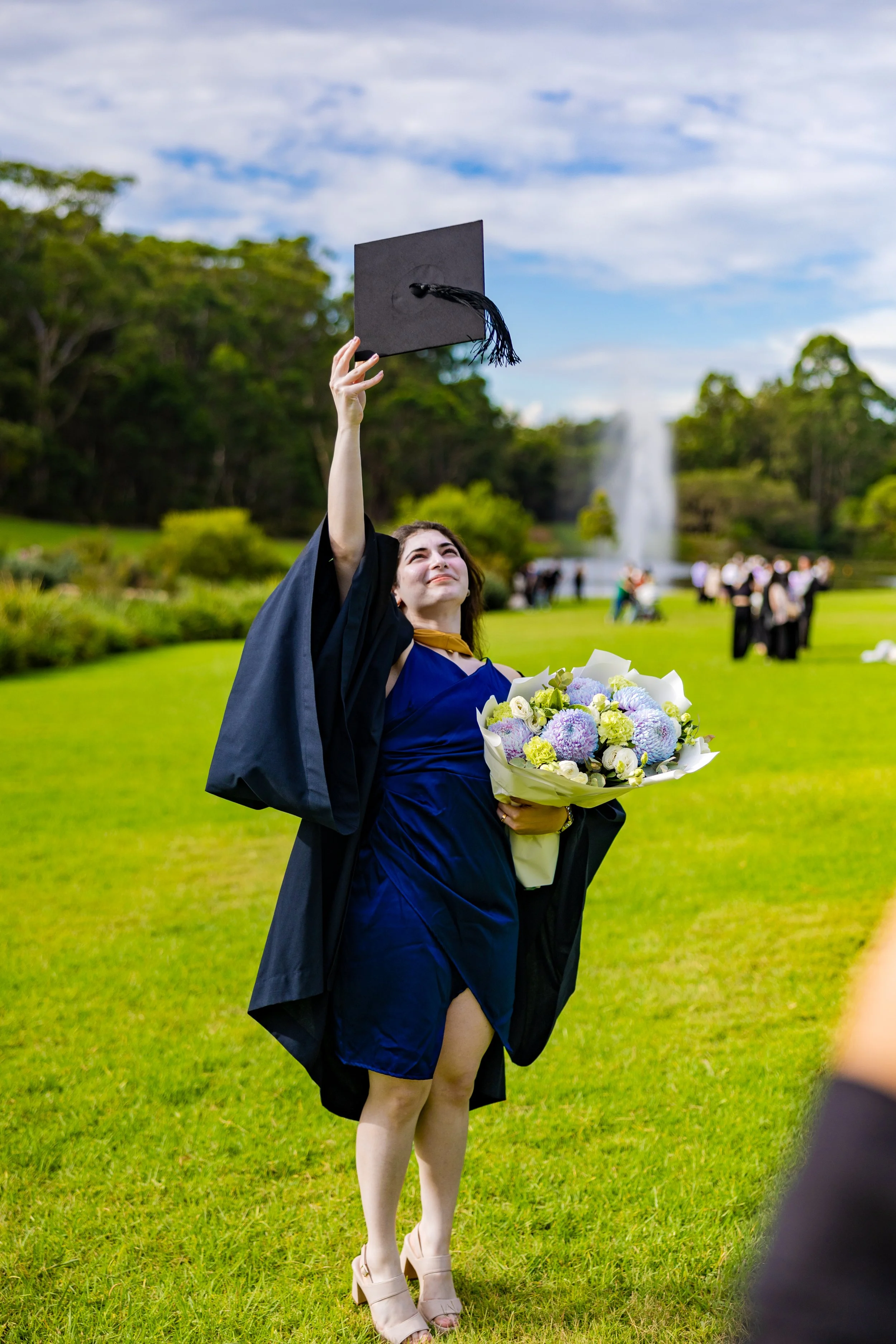A woman in a blue dress and black graduation gown holding a bouquet of flowers and tossing a black graduation cap in the air during a graduation ceremony outdoors.