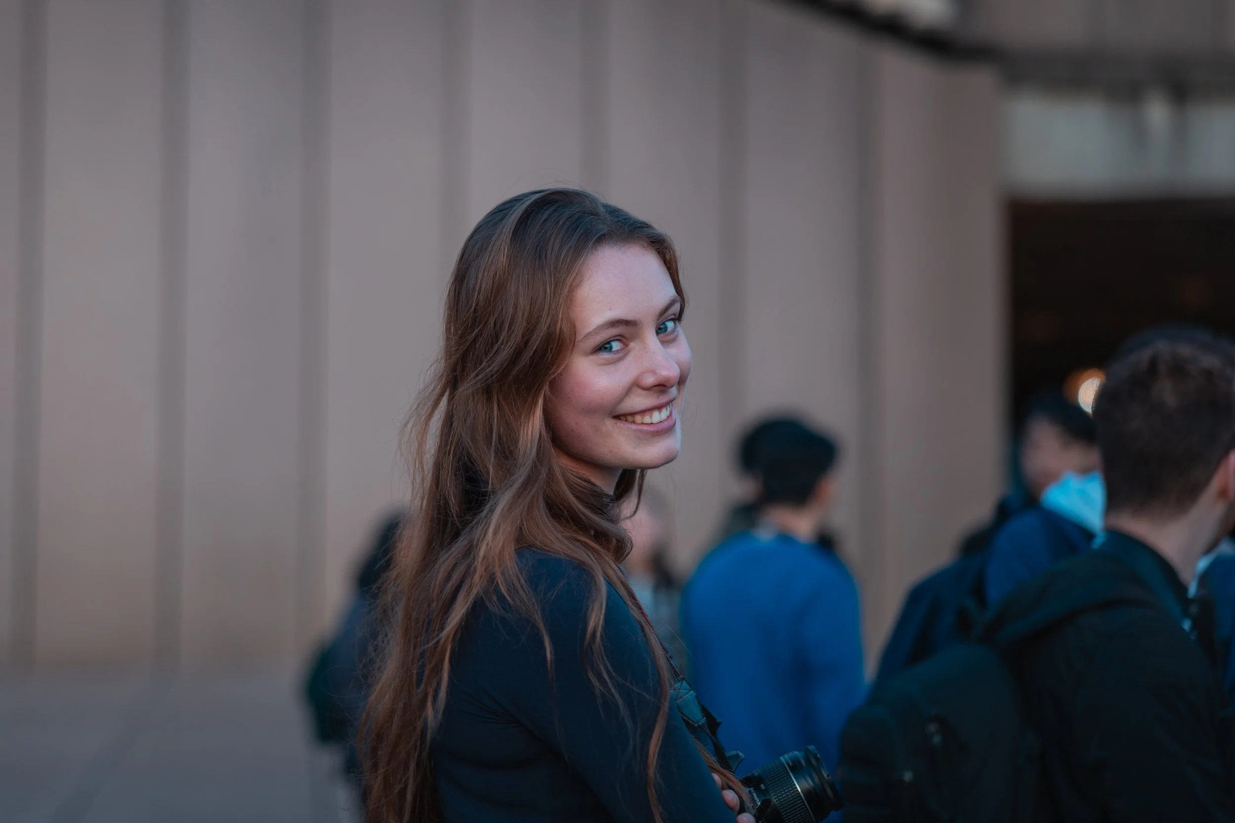 A young woman with long, wavy red hair smiling at the camera, standing outdoors among a group of people.