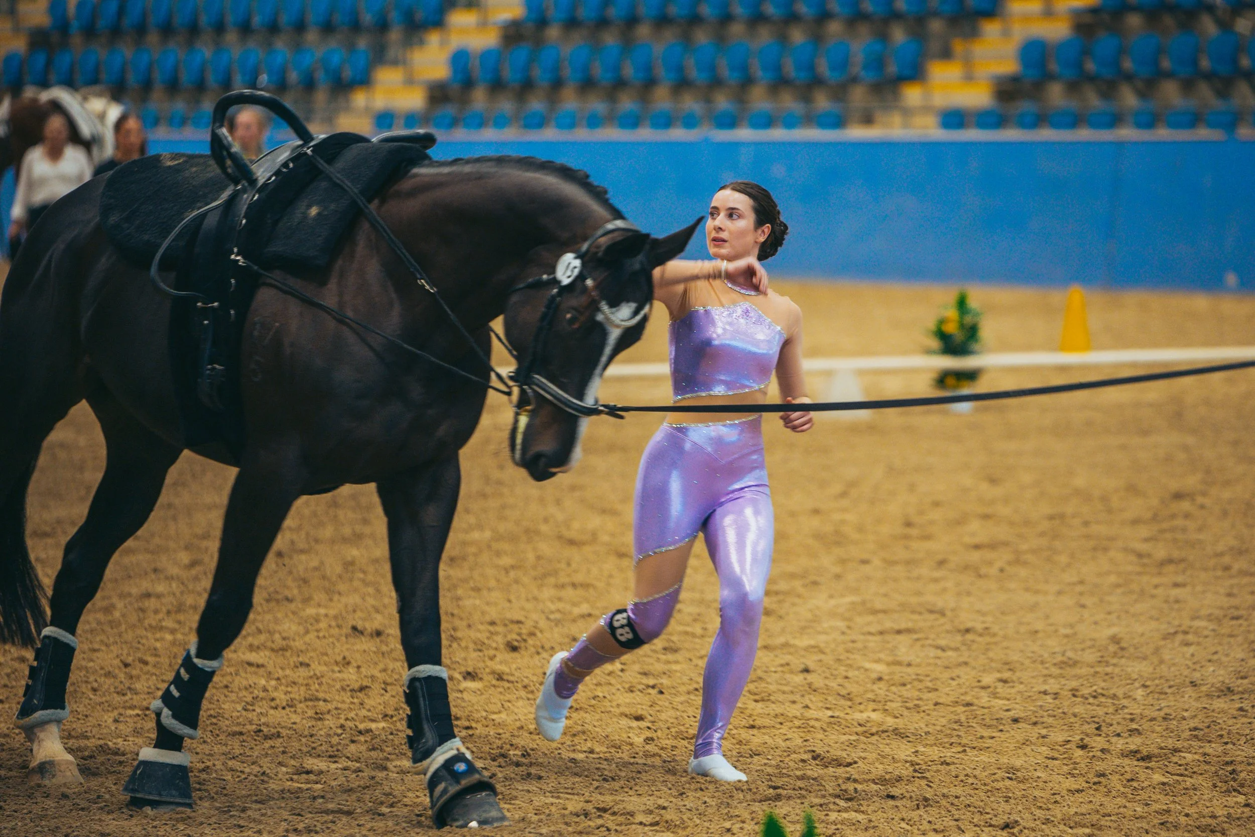 A young woman in an iridescent purple and pink athletic outfit with a knee brace is running alongside a black horse in an indoor arena with empty blue and yellow seats.