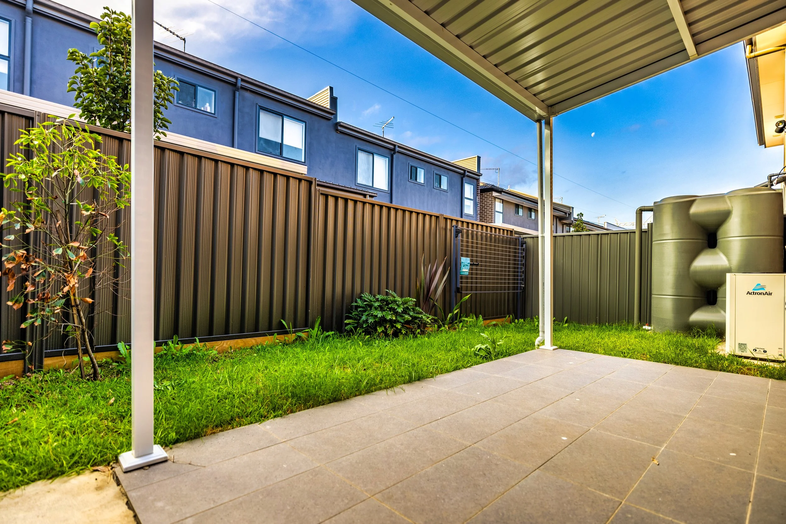 View of a backyard patio area with tiled flooring, a green lawn, plants, and a tall brown fence. There is a covered patio with support beams, a water tank, and an outdoor unit on the side. Residential buildings are visible in the background under a b