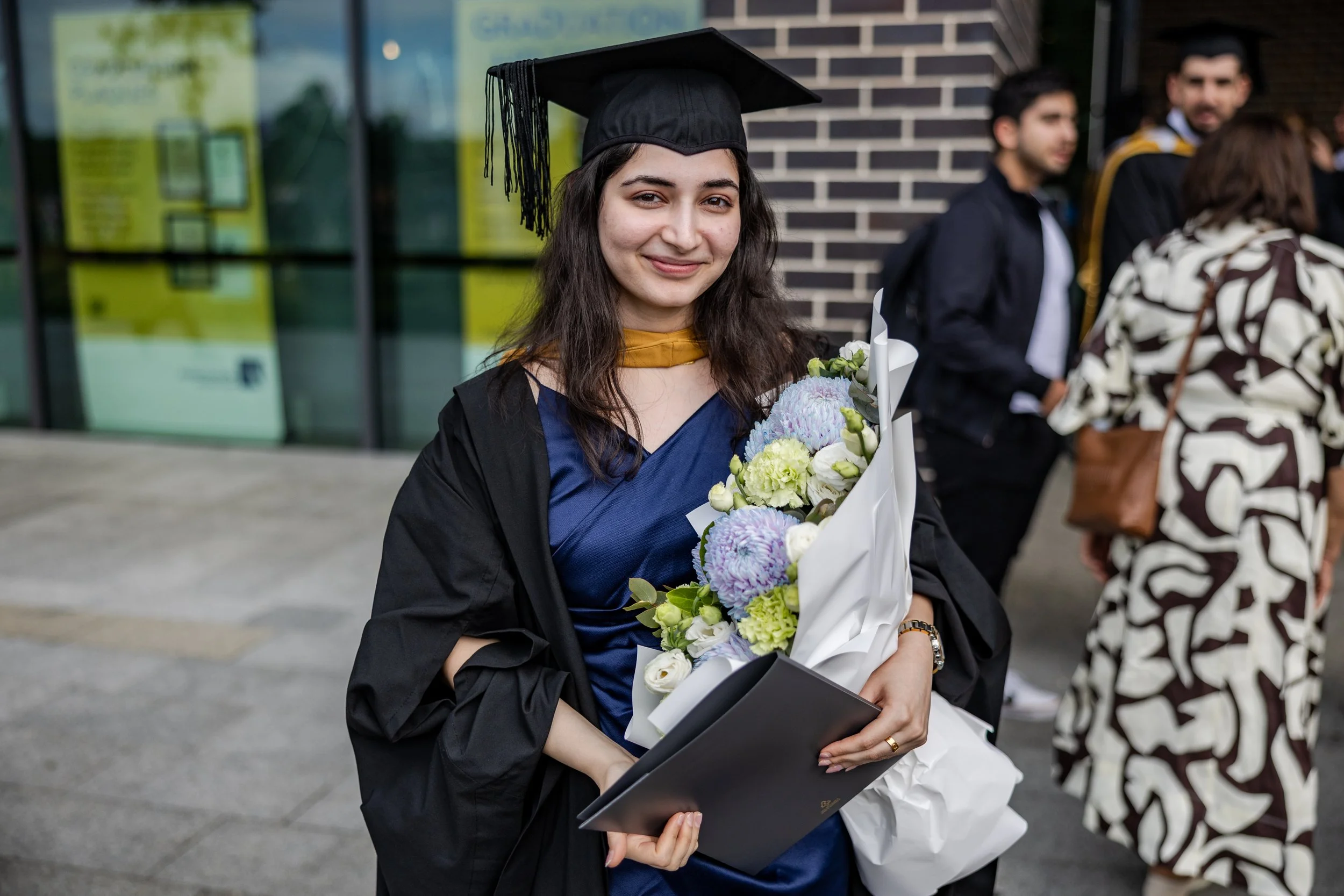 Young woman in graduation cap and gown holding a bouquet of flowers and diploma, smiling outdoors during graduation ceremony.