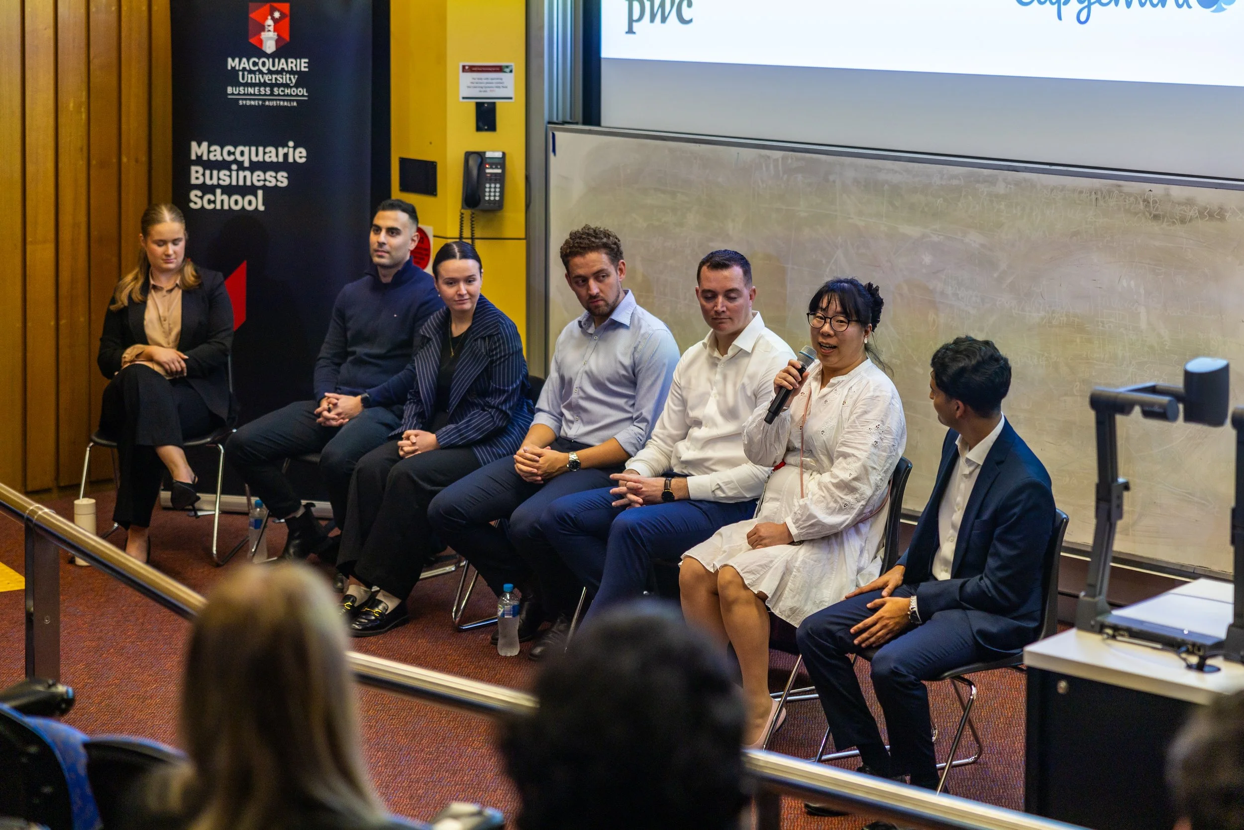 Panel discussion at Macquarie University Business School with seven people seated on stage, one woman speaking into a microphone, audience members in foreground, wall with banners, part of a television screen.