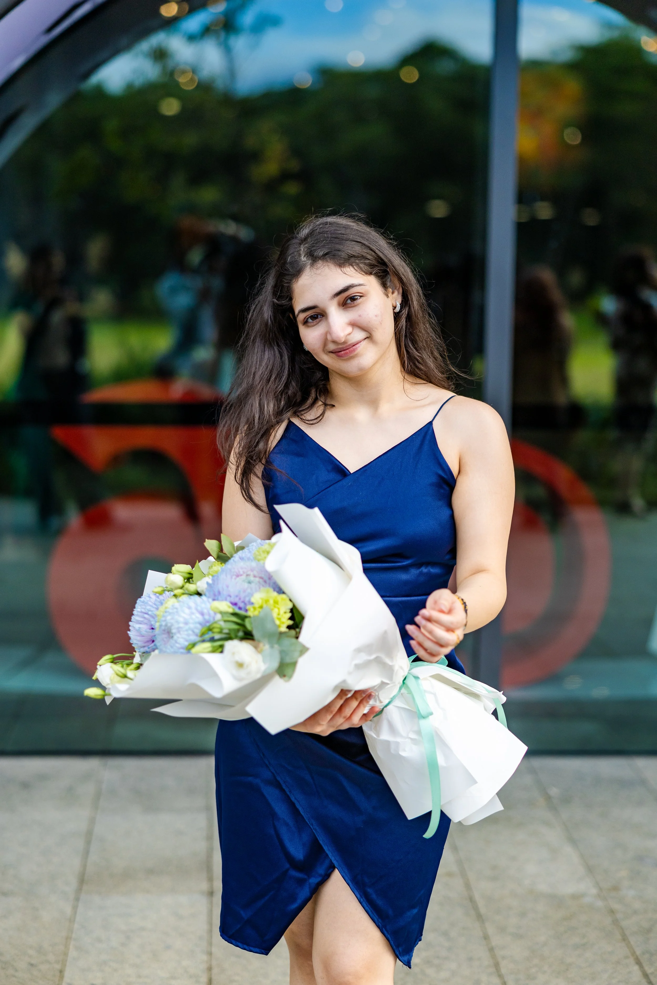 Young woman in a navy blue dress holding a bouquet of flowers, smiling outdoors.