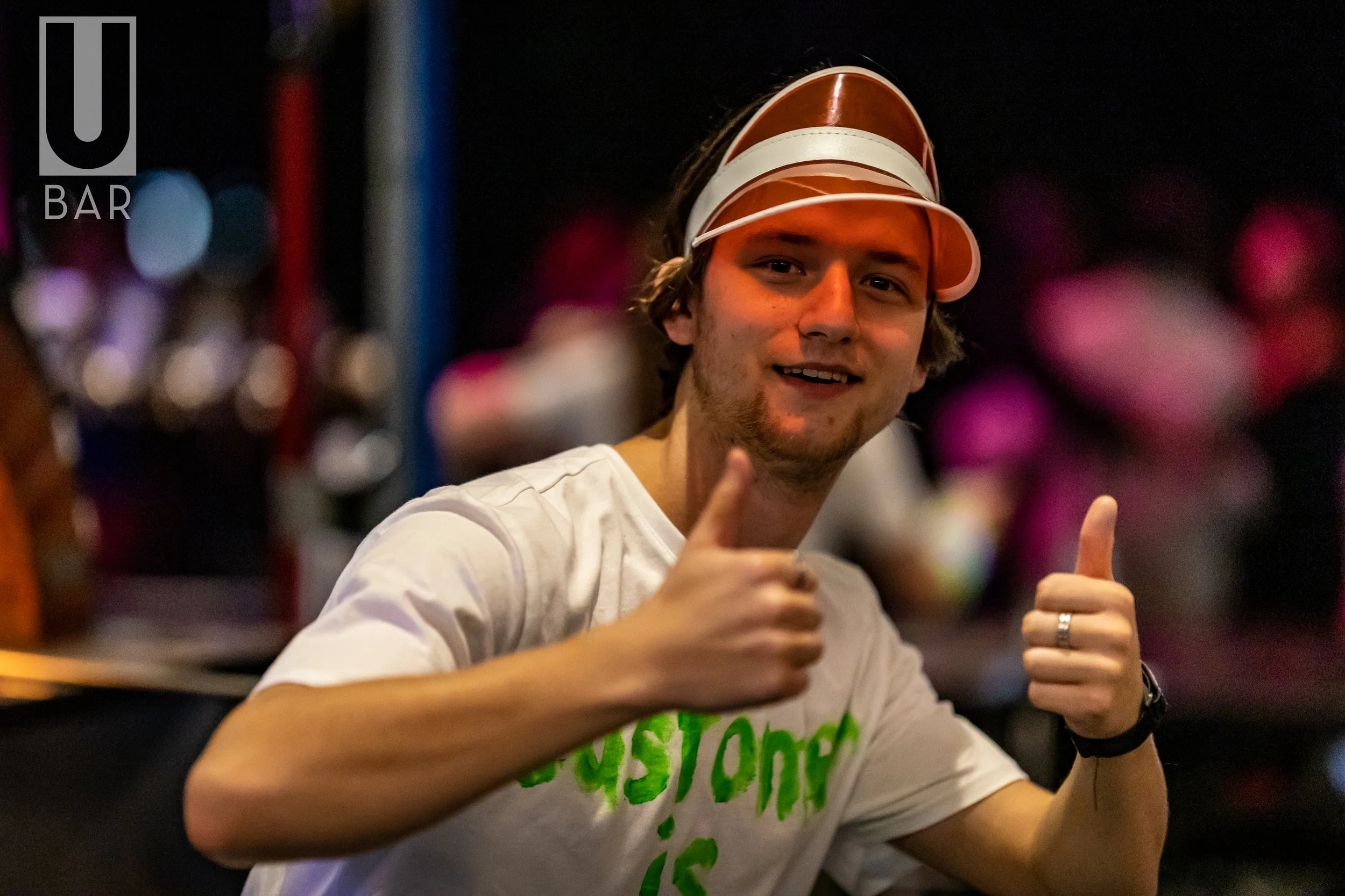 A young man giving two thumbs up inside a bar, wearing a white t-shirt with green writing and a brown and white visor hat.