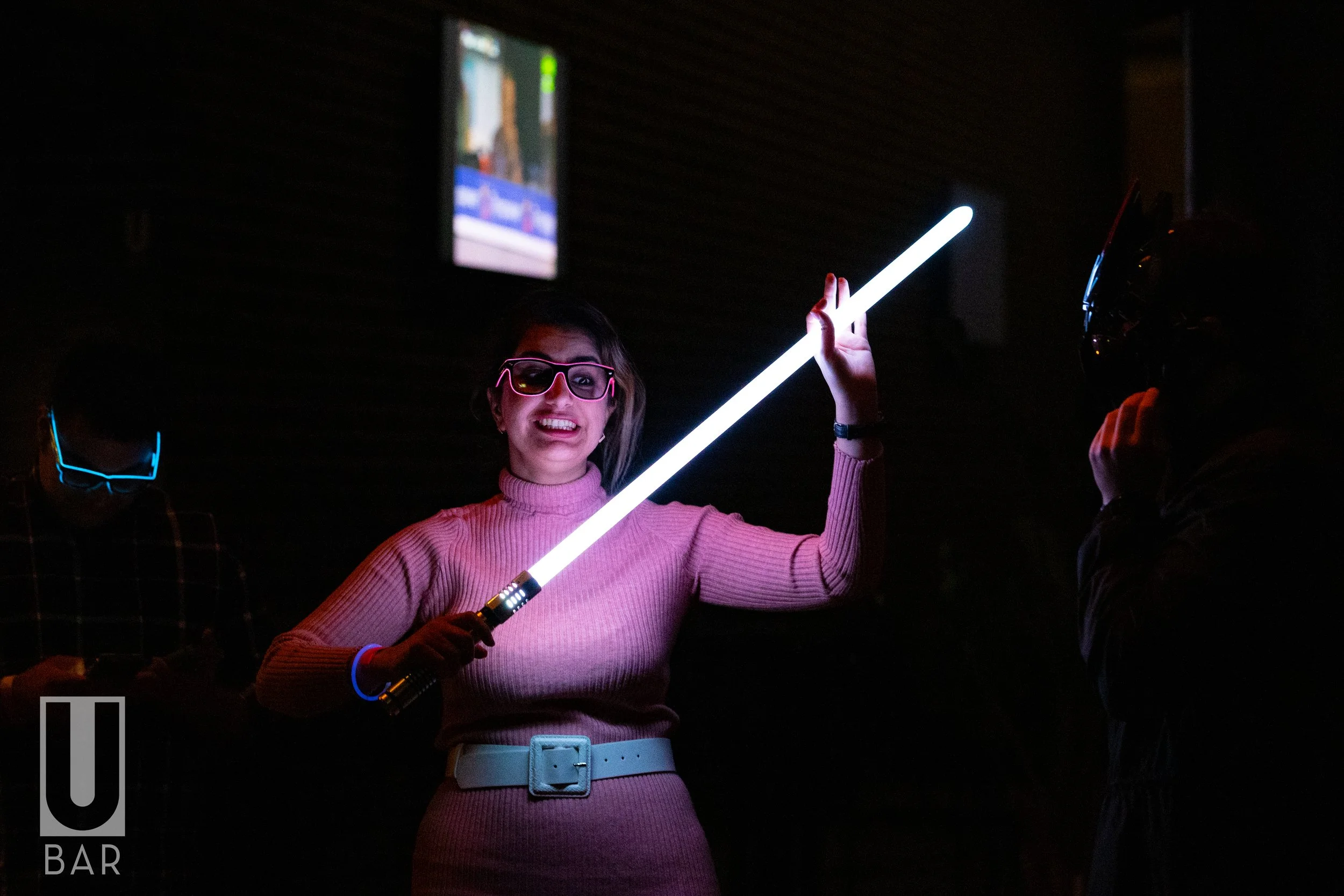 Women with pink sweater and glasses playing with a glowing lightsaber in a dark bar with a TV screen in the background.