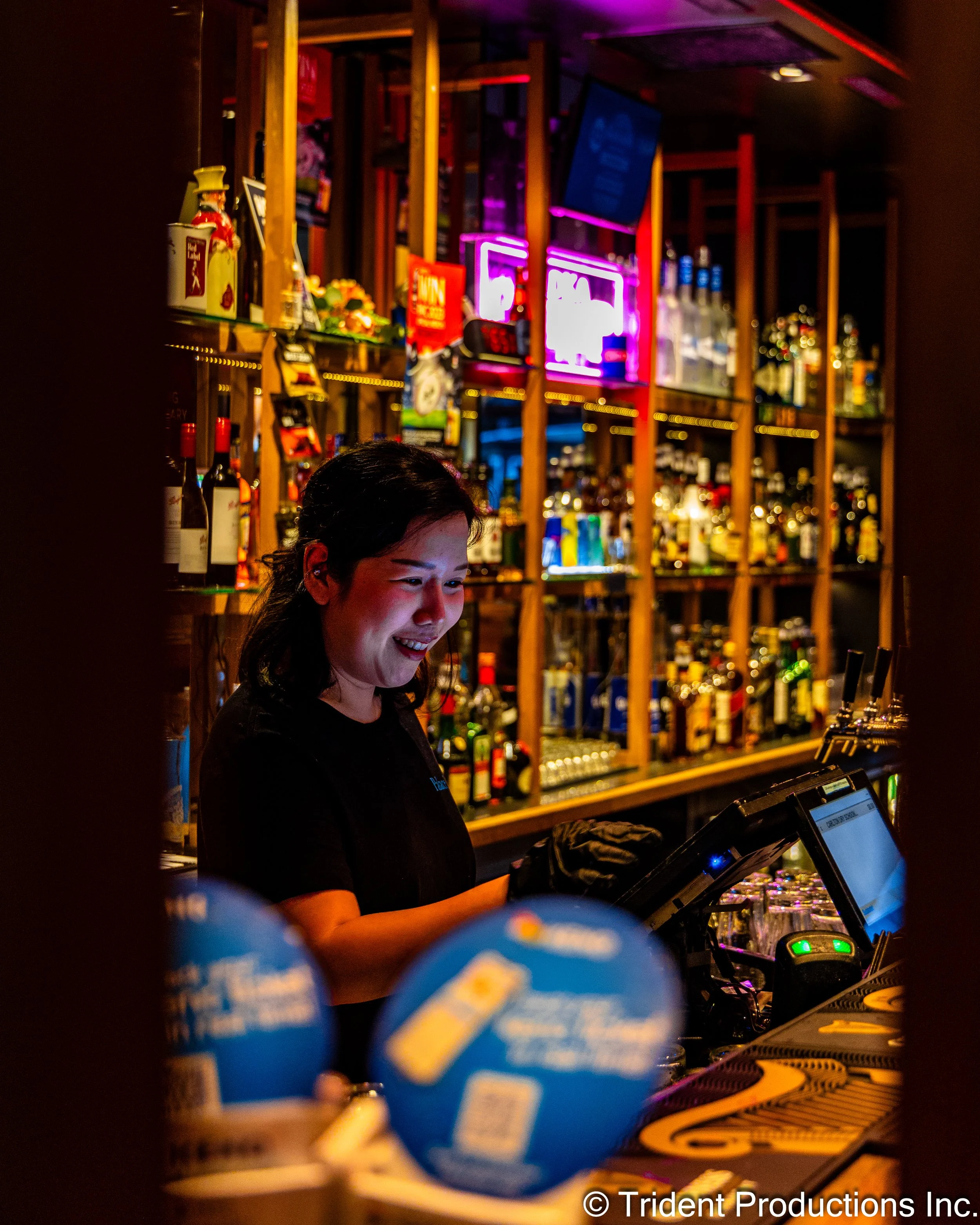 A female bartender smiling while working behind a bar with shelves of liquor bottles, neon signs, and a cash register.