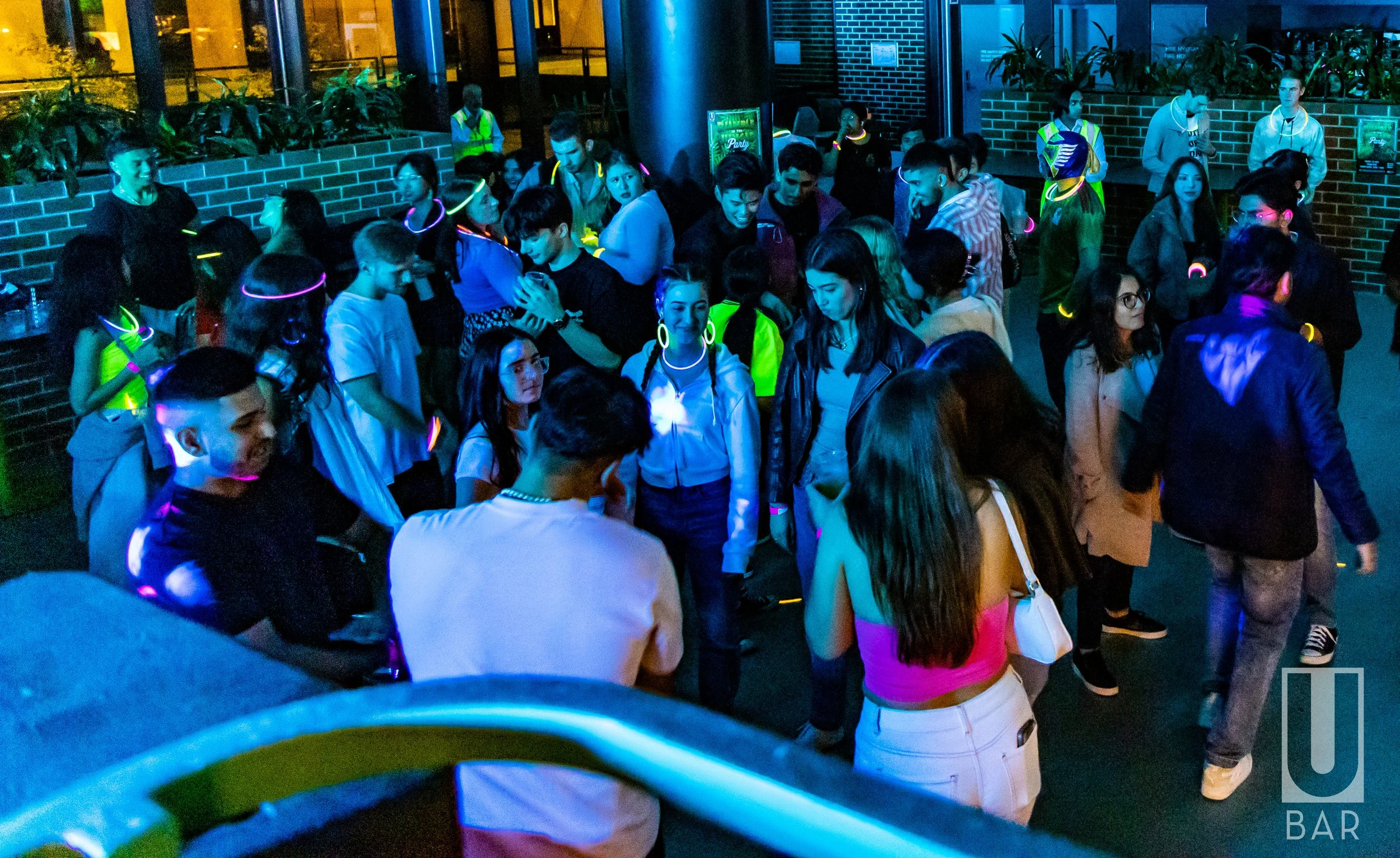 A group of young people at a party or nightclub with glow-in-the-dark jewelry and accessories, standing outside near a brick wall and plants, with blue and yellow lighting.