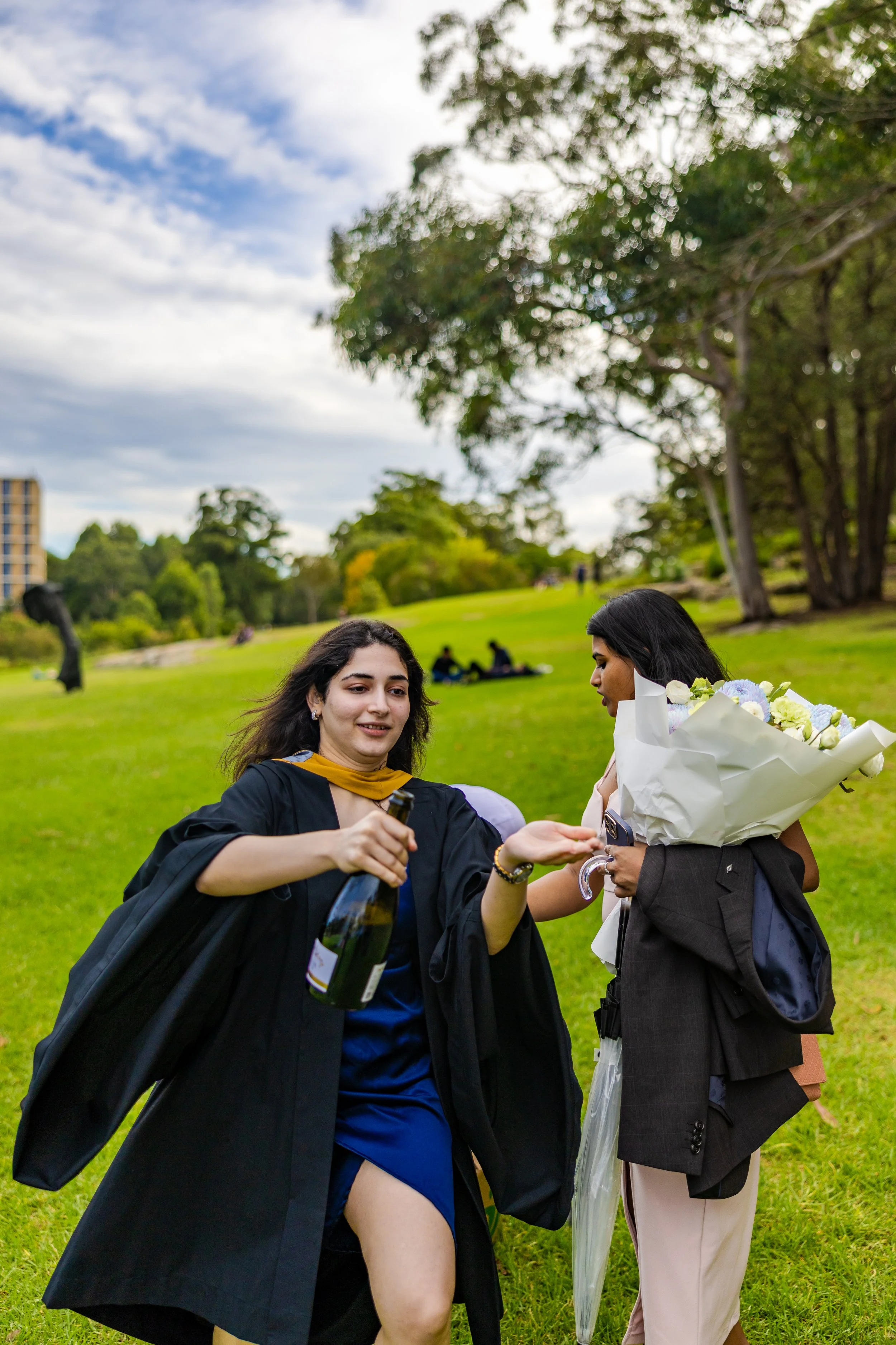 Two women celebrating outdoors, one in a graduation gown holding a bottle, the other in business attire holding a bouquet of flowers and a folded umbrella, on a grassy field with trees and cloudy sky in the background.