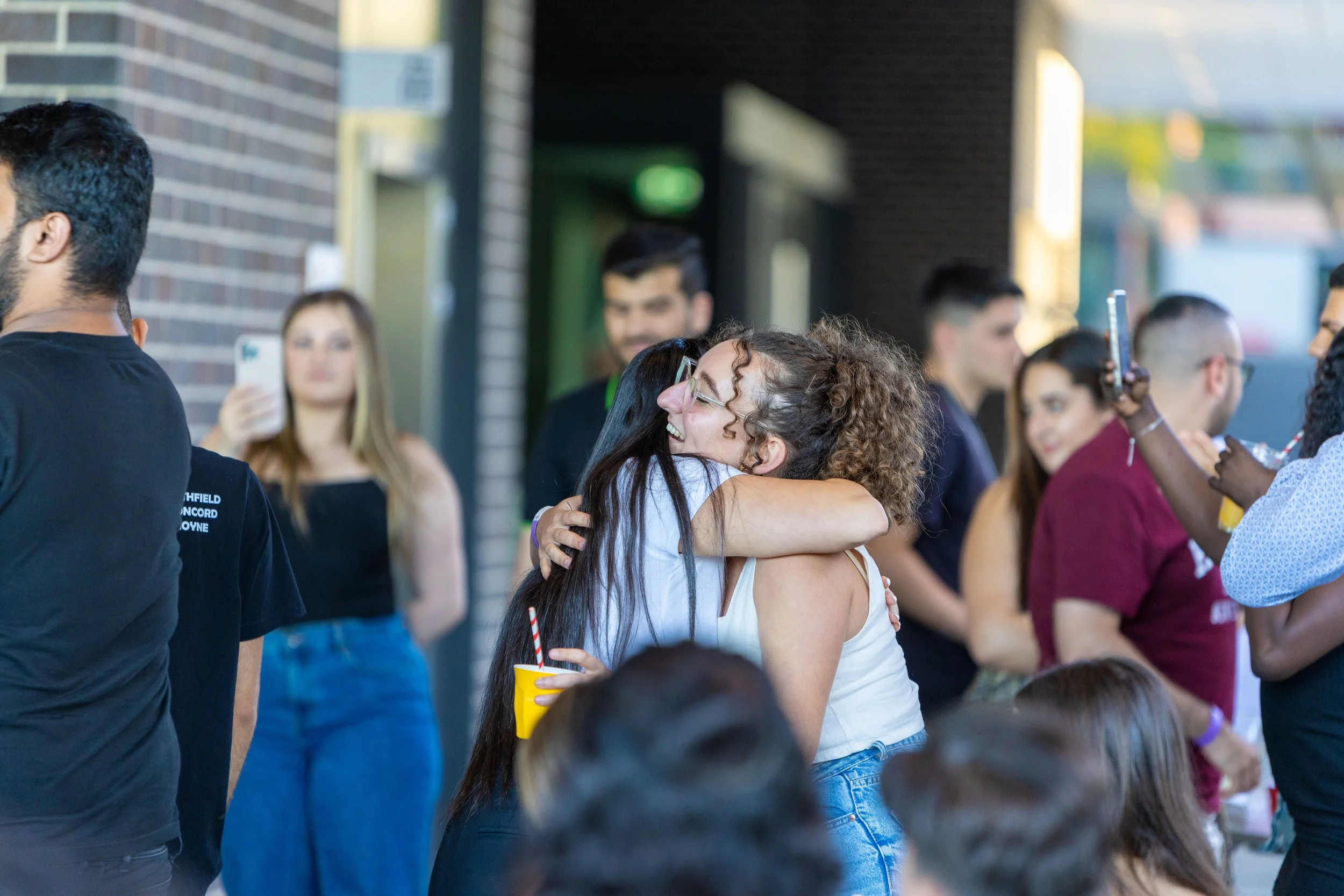 Two women hugging each other at an outdoor gathering, with several other people in the background, some taking photos and chatting.