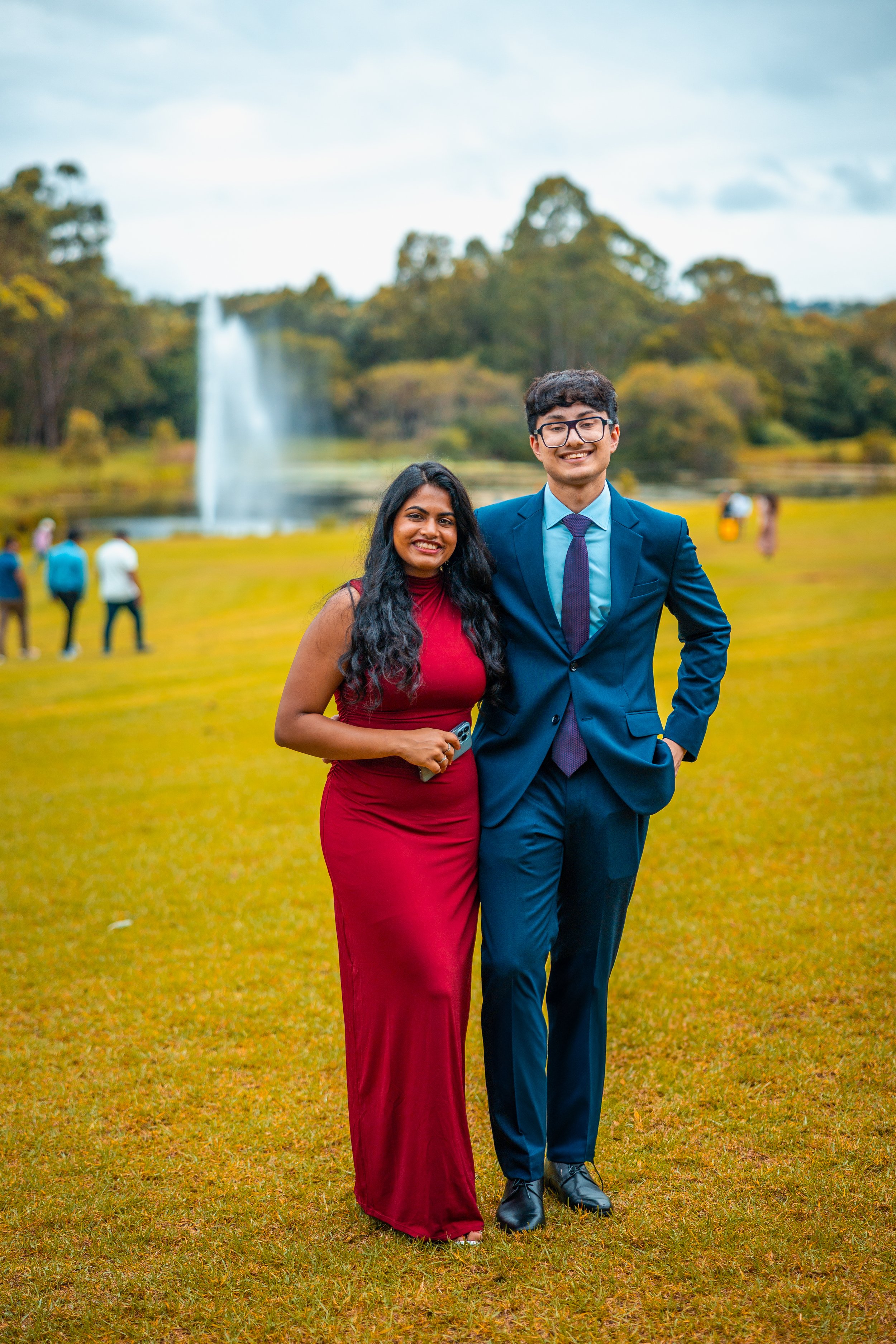 A smiling woman in a red dress and a smiling man in a blue suit standing close together outdoors on grass, with a waterfall and trees in the background, and other people in the distance.