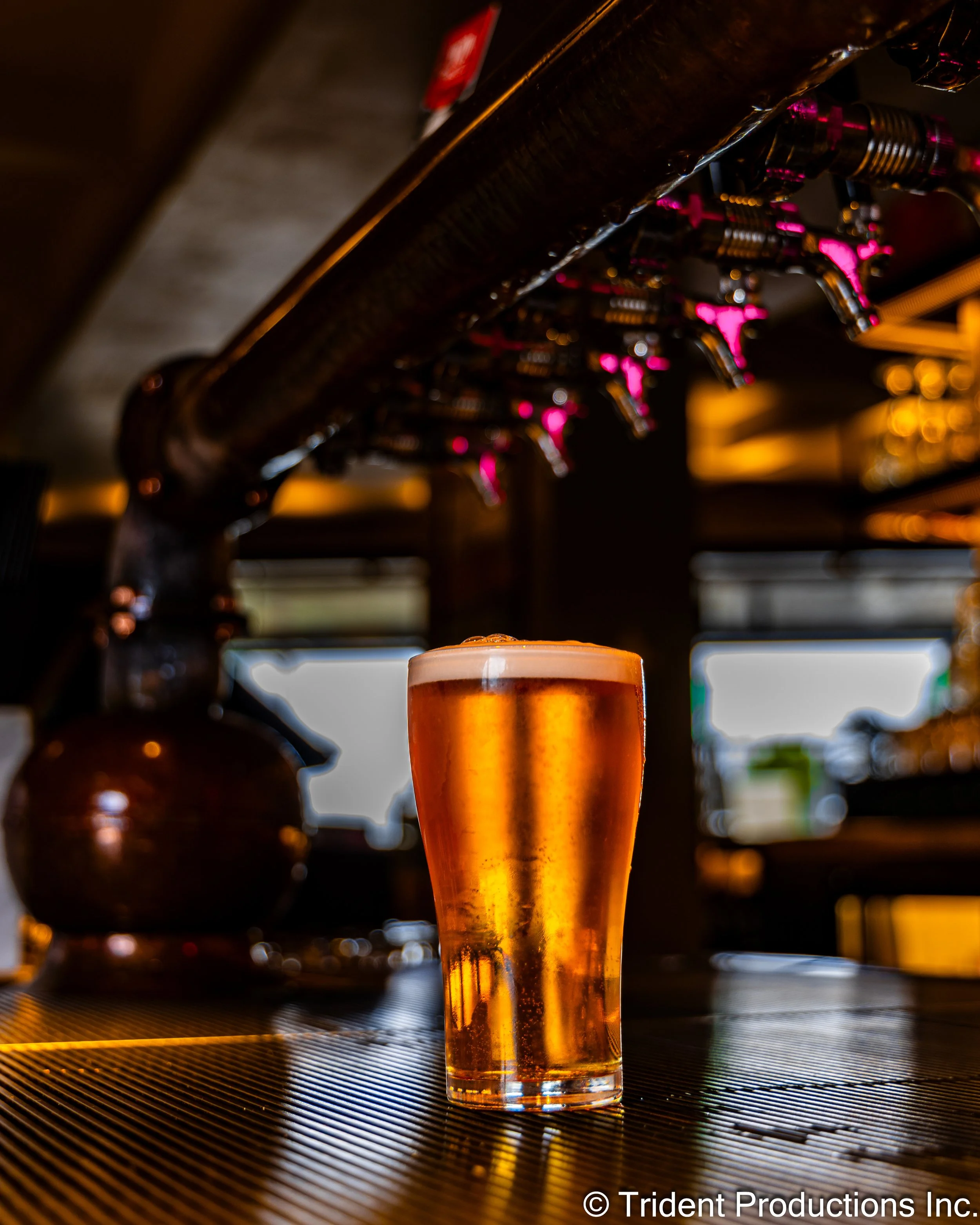 Pint of amber beer with foam on top on a bar counter under beer taps in a dimly lit pub.