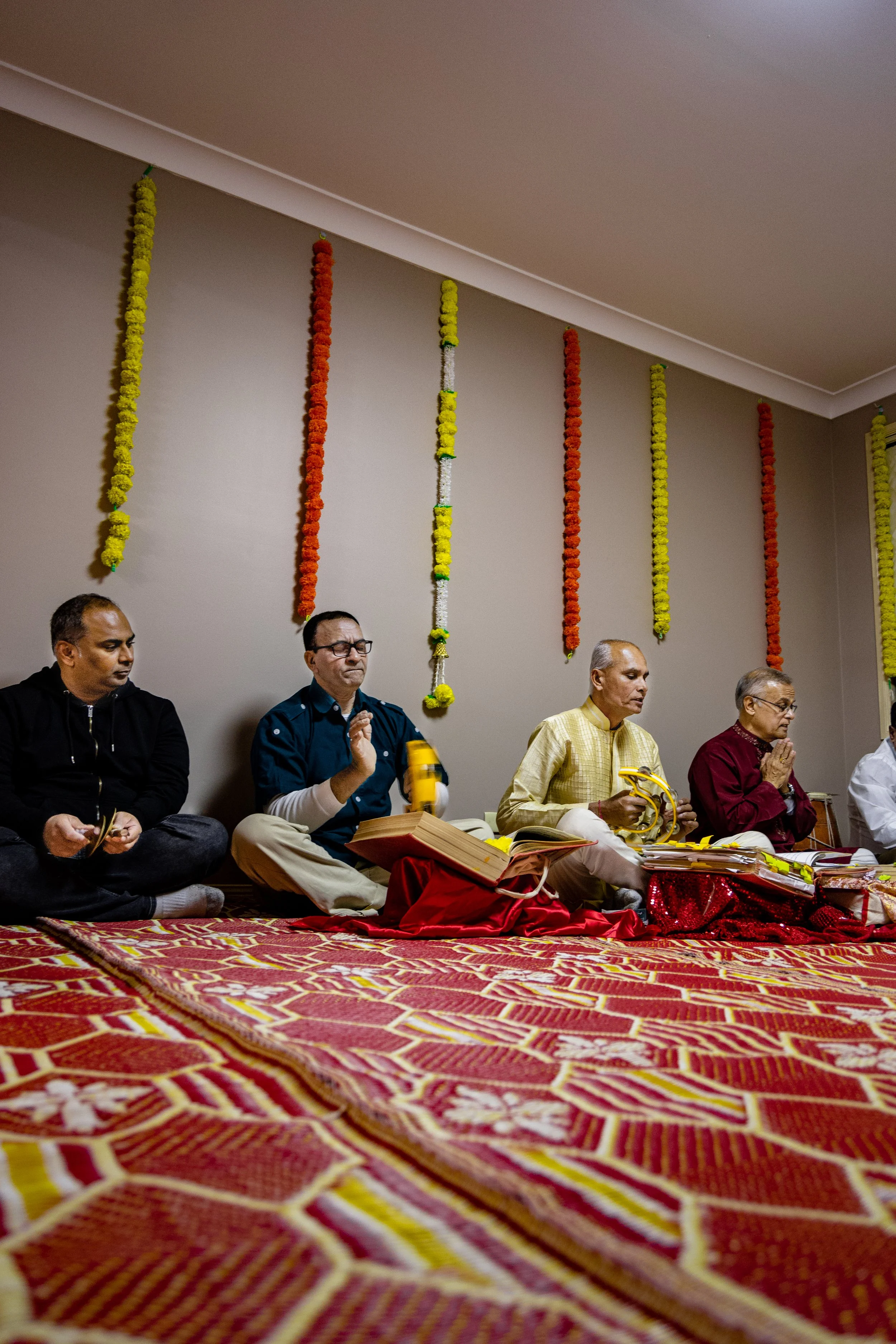 A group of men participating in a traditional Indian religious ceremony, sitting cross-legged on a decorated carpet with offerings and scriptures, with floral garlands hanging on the wall behind them.
