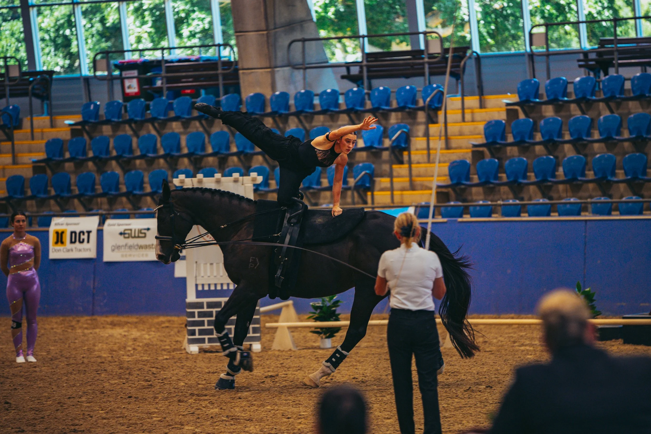 A female equestrian performer balances on a black horse inside an indoor arena, with an audience in the background.