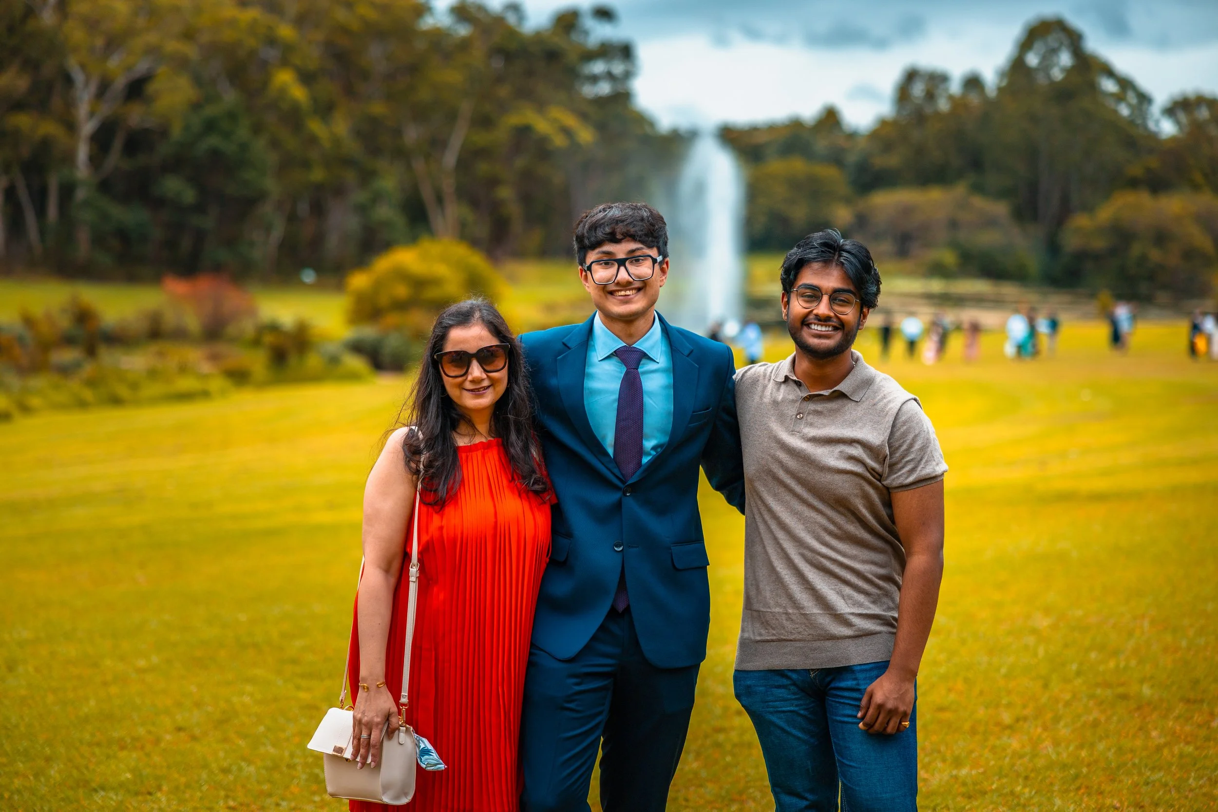 Three people standing in a park with trees and a fountain in the background. The woman on the left is wearing a red dress and sunglasses, the man in the middle is in a blue suit, and the man on the right is in a gray shirt and glasses. They are smili