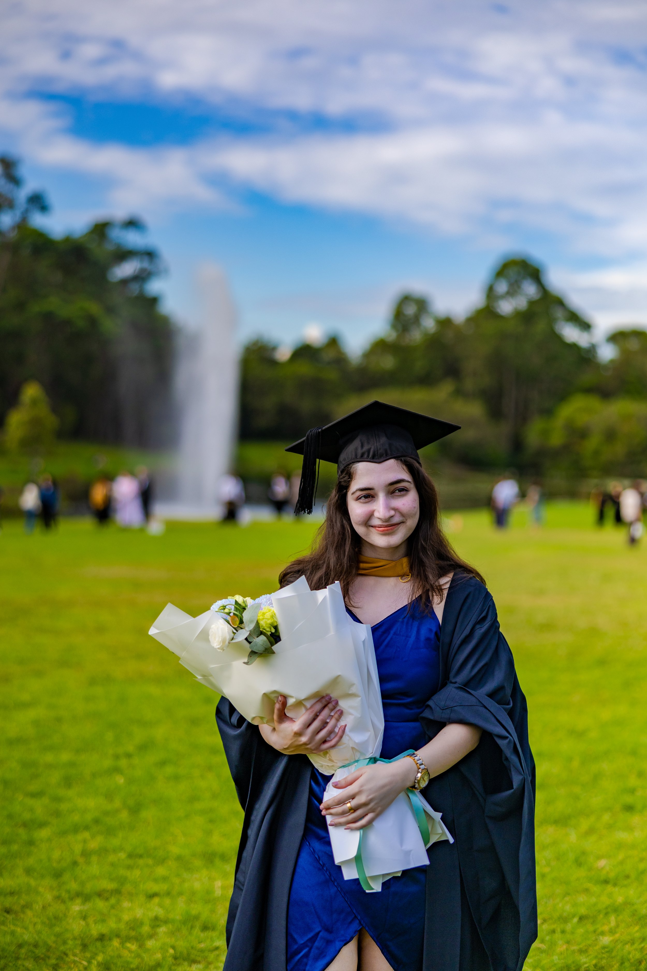A young woman in a graduation cap and gown holding a bouquet of white flowers outdoors with a fountain and trees in the background.