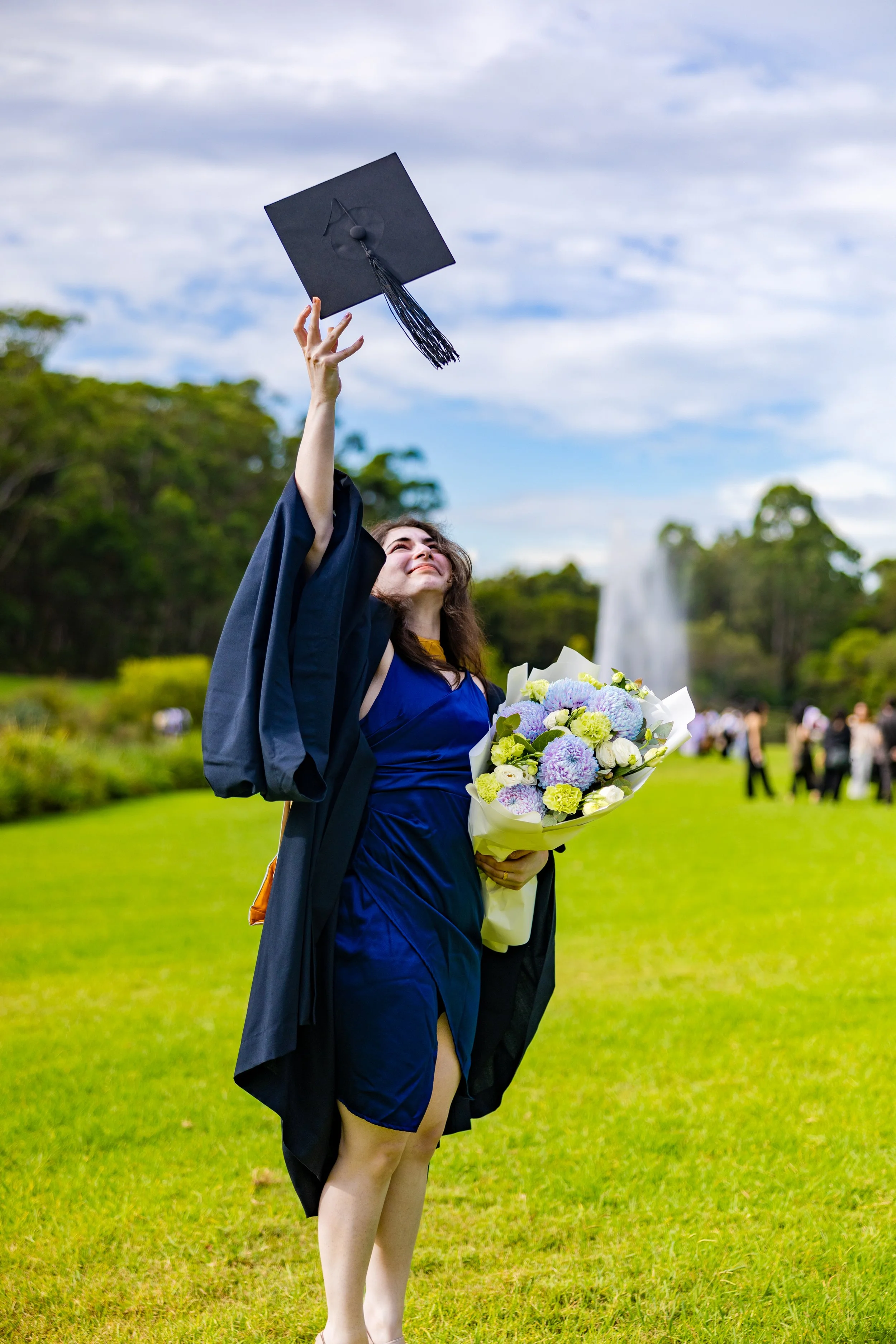 A woman in a blue dress and graduation gown holding a bouquet of flowers, celebrating outdoors on graduation day.