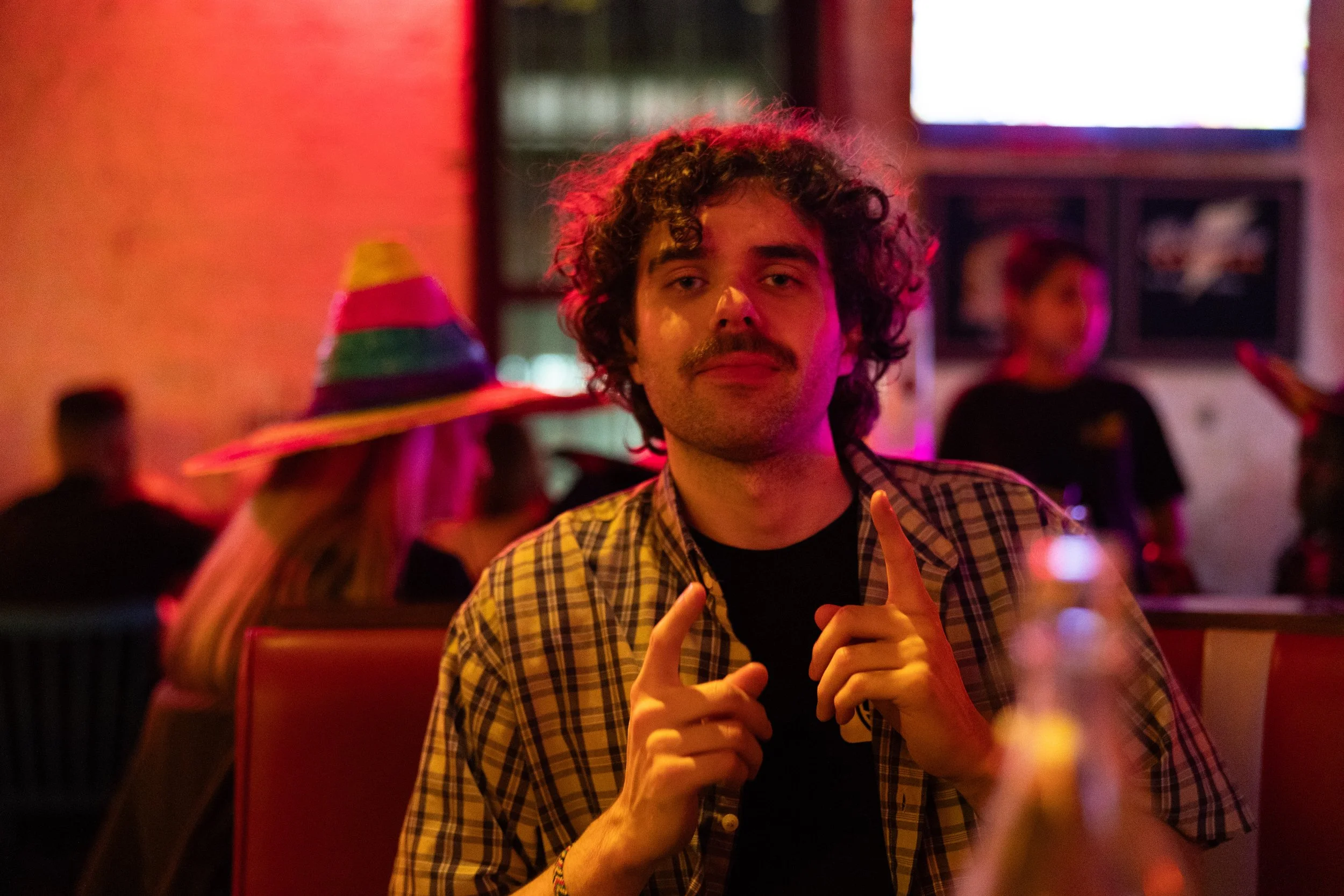 A young man with curly hair and a mustache, wearing a checkered shirt, sitting in a dimly lit restaurant or bar with pink and purple lighting, pointing upwards with both index fingers.
