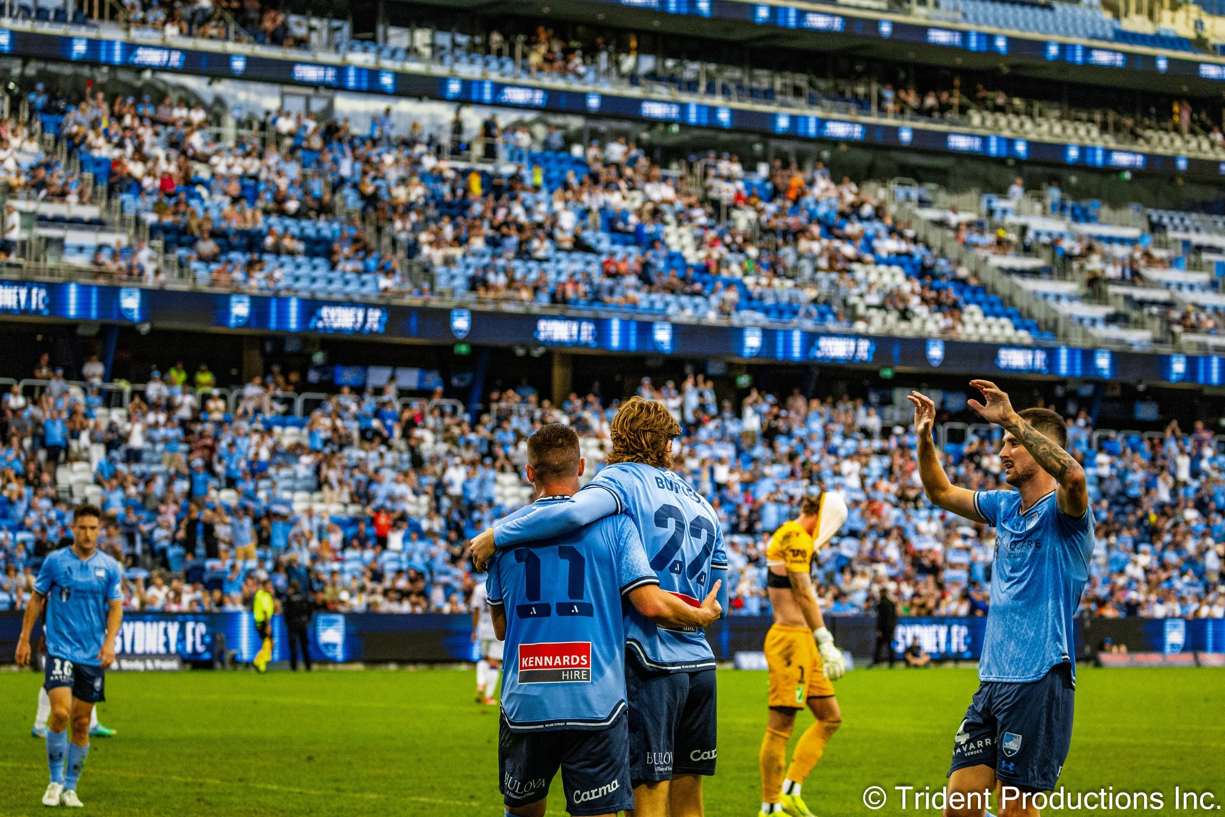 Soccer players in blue uniforms celebrating on the field, with one player giving a hug to a teammate, a player in a yellow goalkeeper uniform in the background, and a crowd of spectators in the stands.