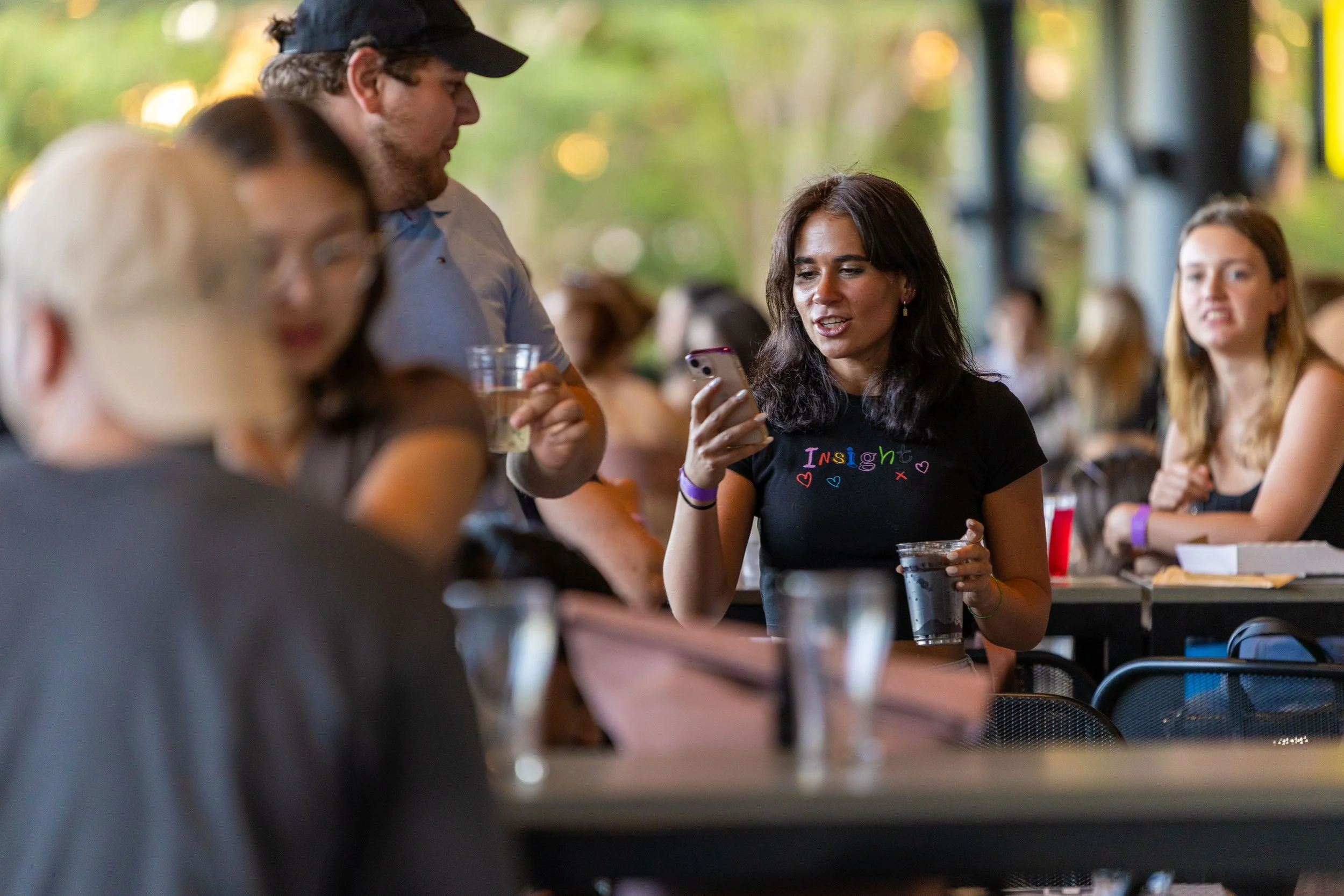 Group of people socializing at a cafe, with a woman showing her phone to others, some holding drinks, in a lively indoor setting with greenery outside.