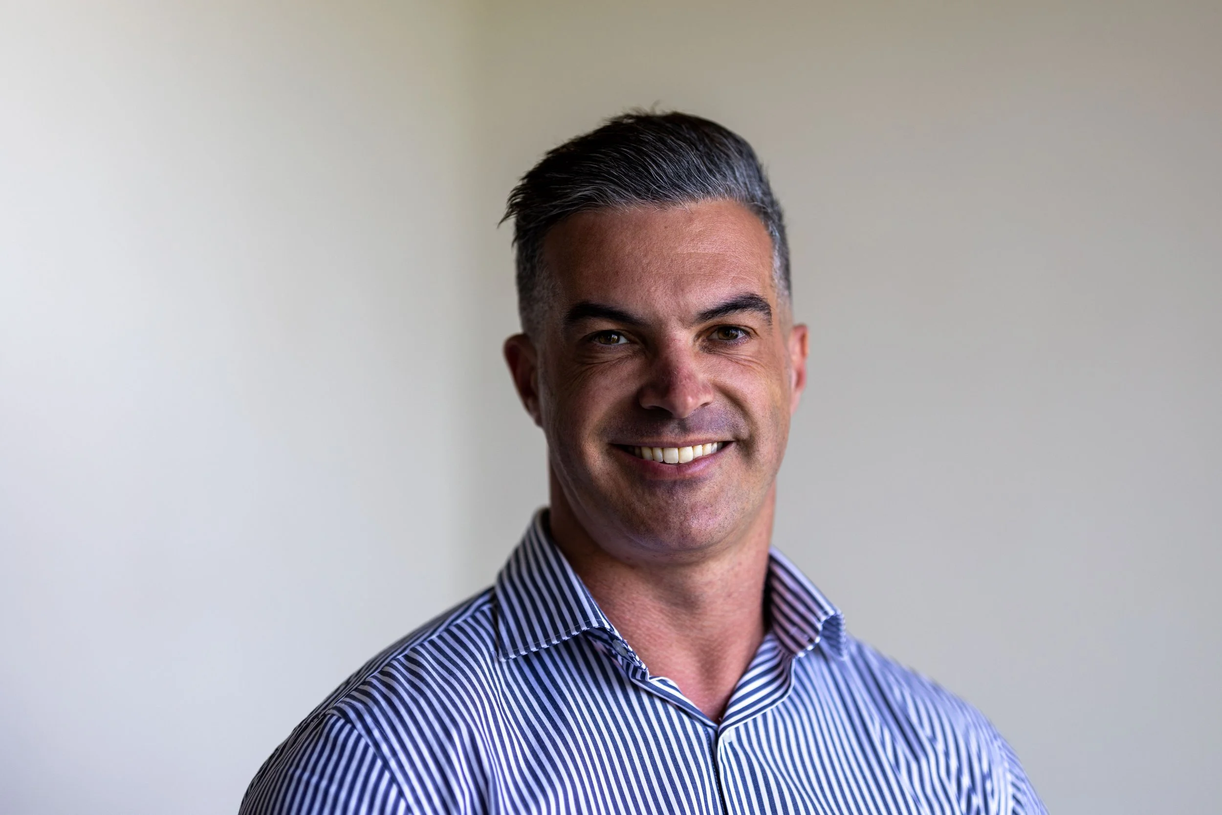Portrait of a smiling man with short dark hair, wearing a navy and white striped dress shirt, standing against a plain off-white wall.