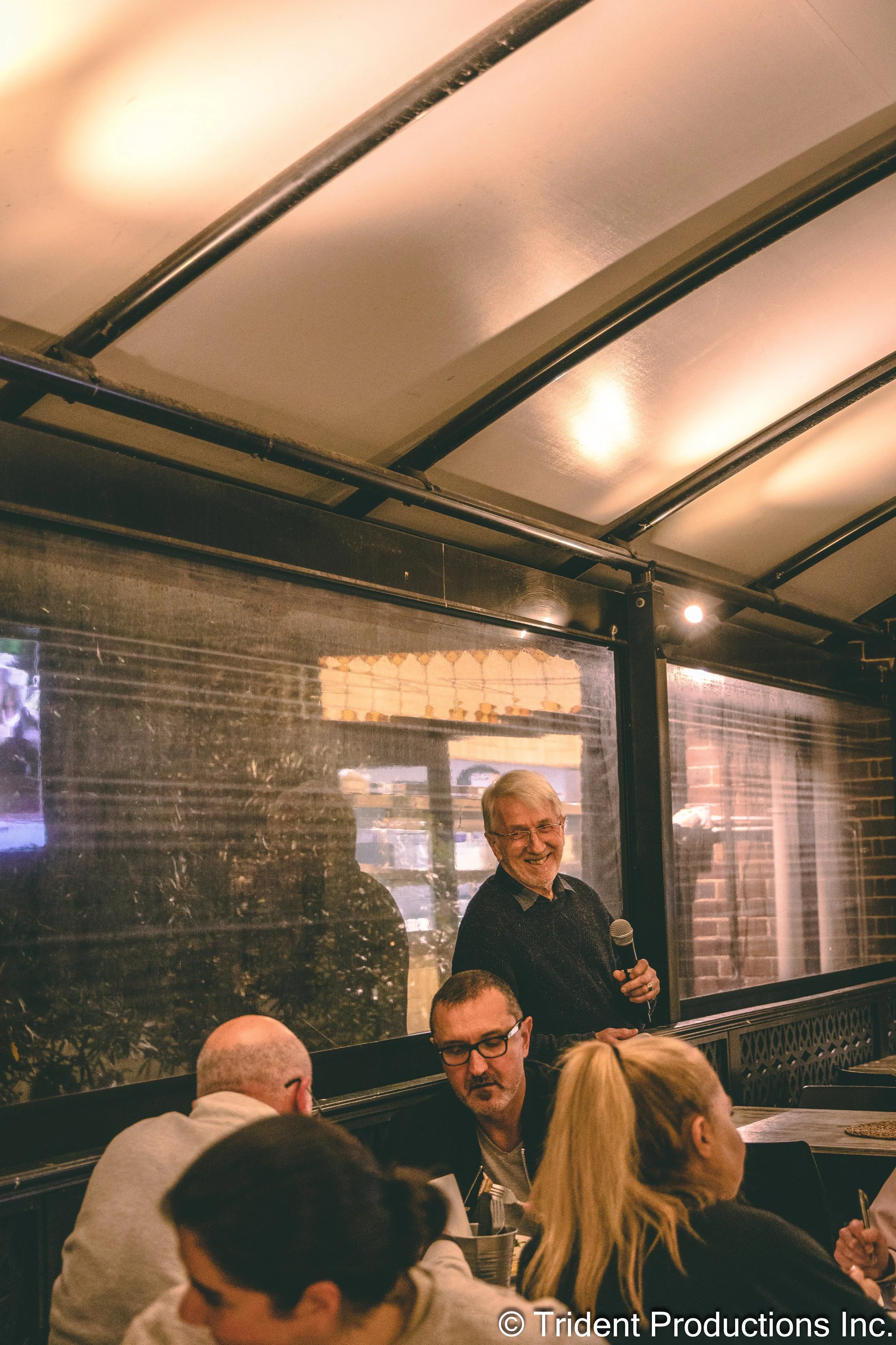 An elderly man standing with a microphone, smiling, while speaking to a group of people in a restaurant or cafe.