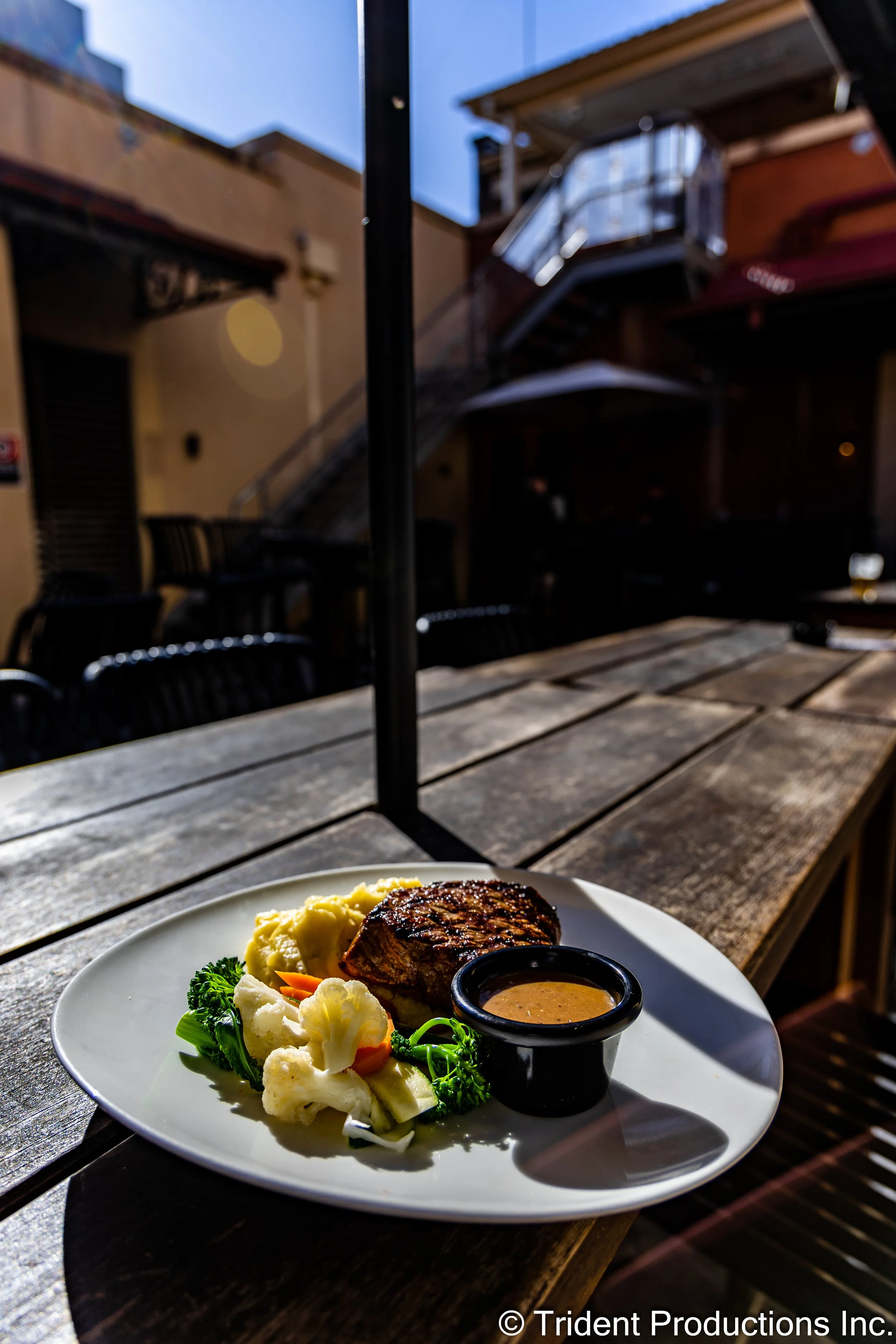Plate of grilled steak with mashed potatoes, steamed vegetables, and gravy sauce on a wooden table outdoors.