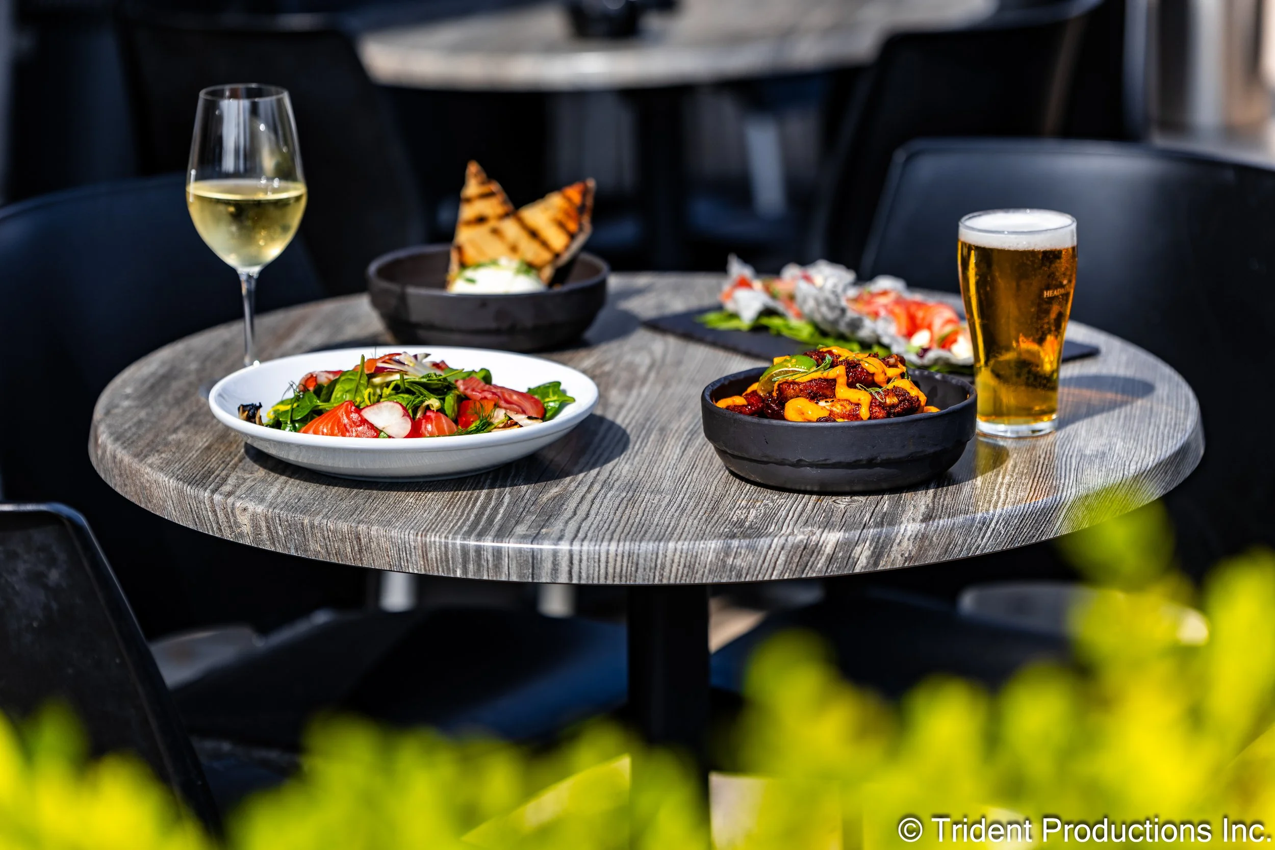 A round outdoor table with a variety of food and drinks including a glass of white wine, a pint of beer, a salad with greens and tomatoes, a bowl of nachos with cheese, a bowl of sushi rolls, and grilled bread.