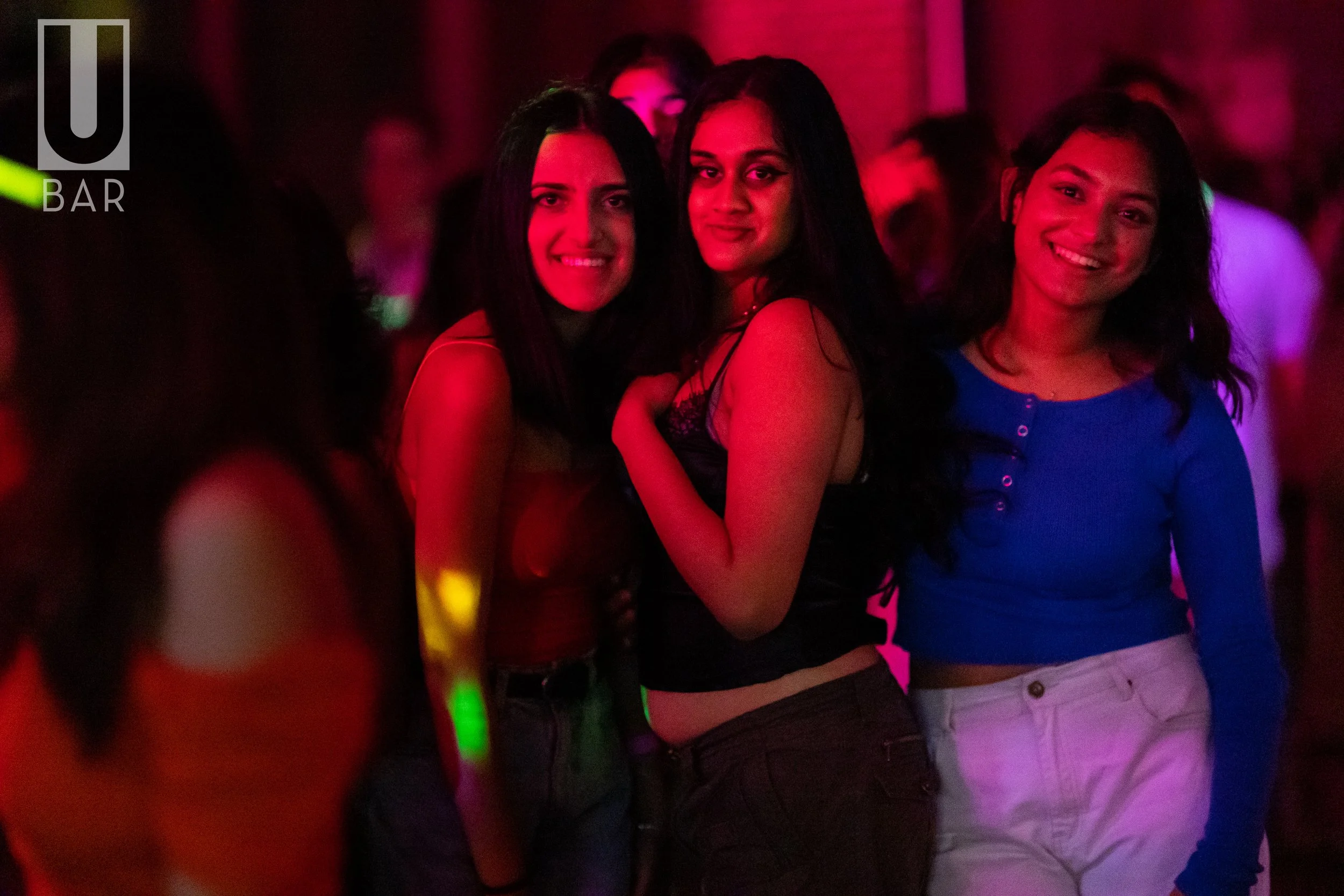Group of three women dancing and smiling at a nightclub with colorful lighting.