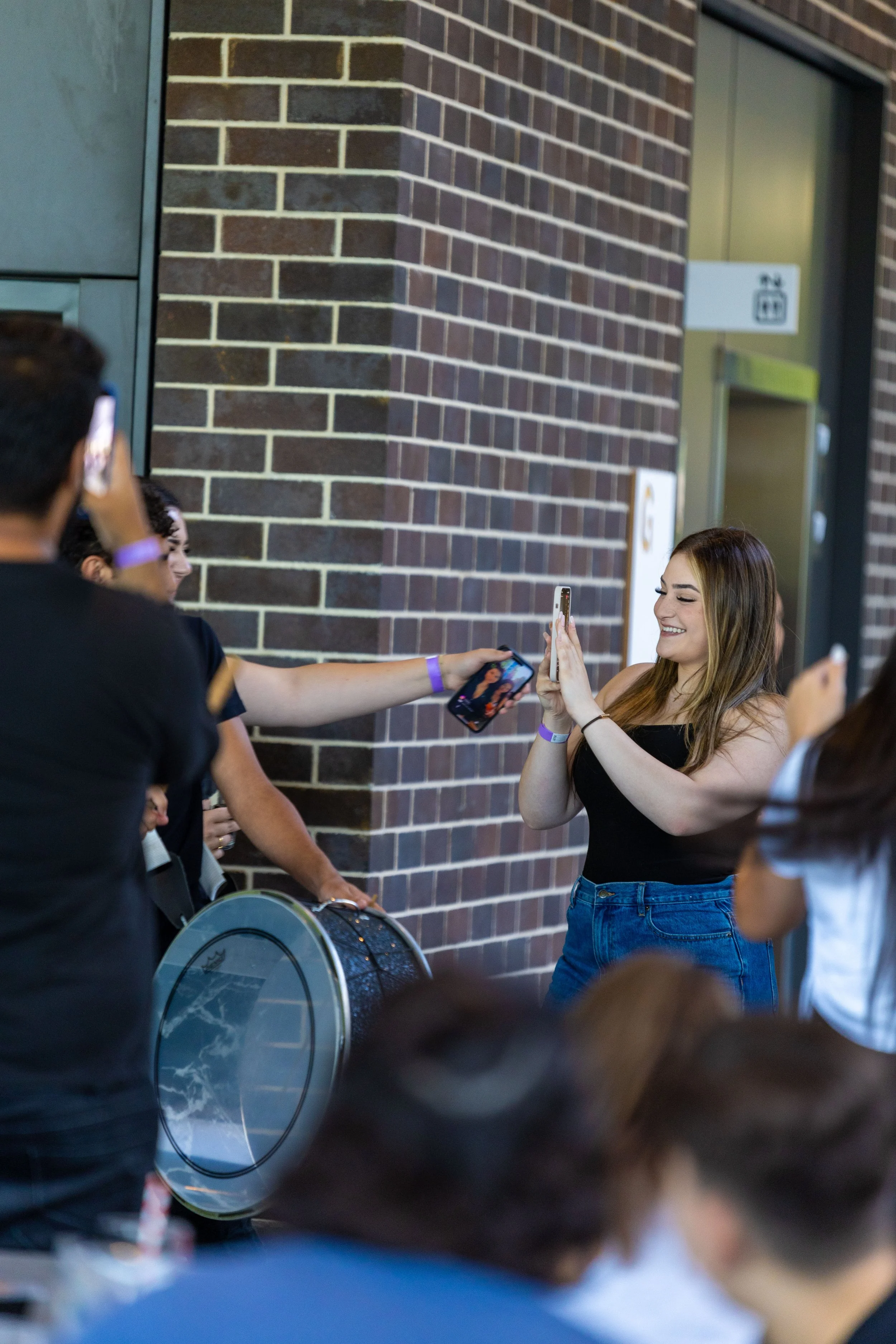 A group of people outdoors near a brick building, with a woman smiling and taking a selfie while others also pose for photos. One person holds a drum.
